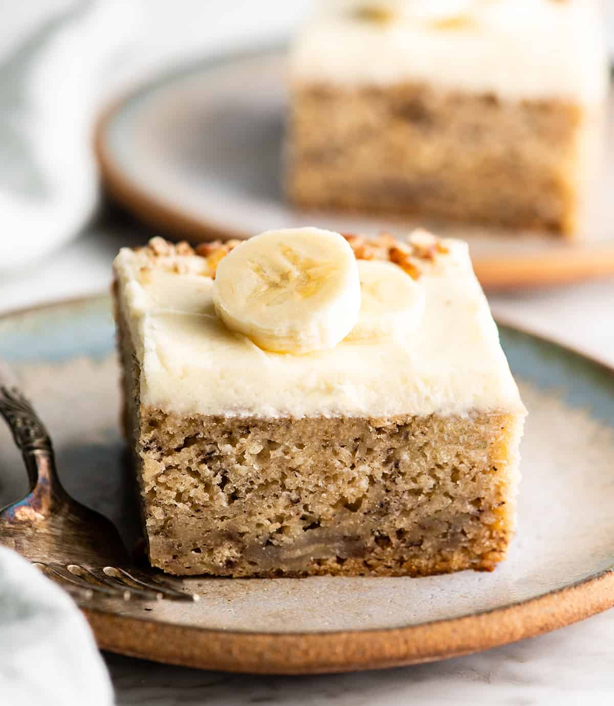 This is the best banana cake recipe ever! It's easy to make (no mixer required), moist, loaded with bananas and topped with a delicious cream cheese frosting. front view of a piece of banana cake with cream cheese frosting on a plate