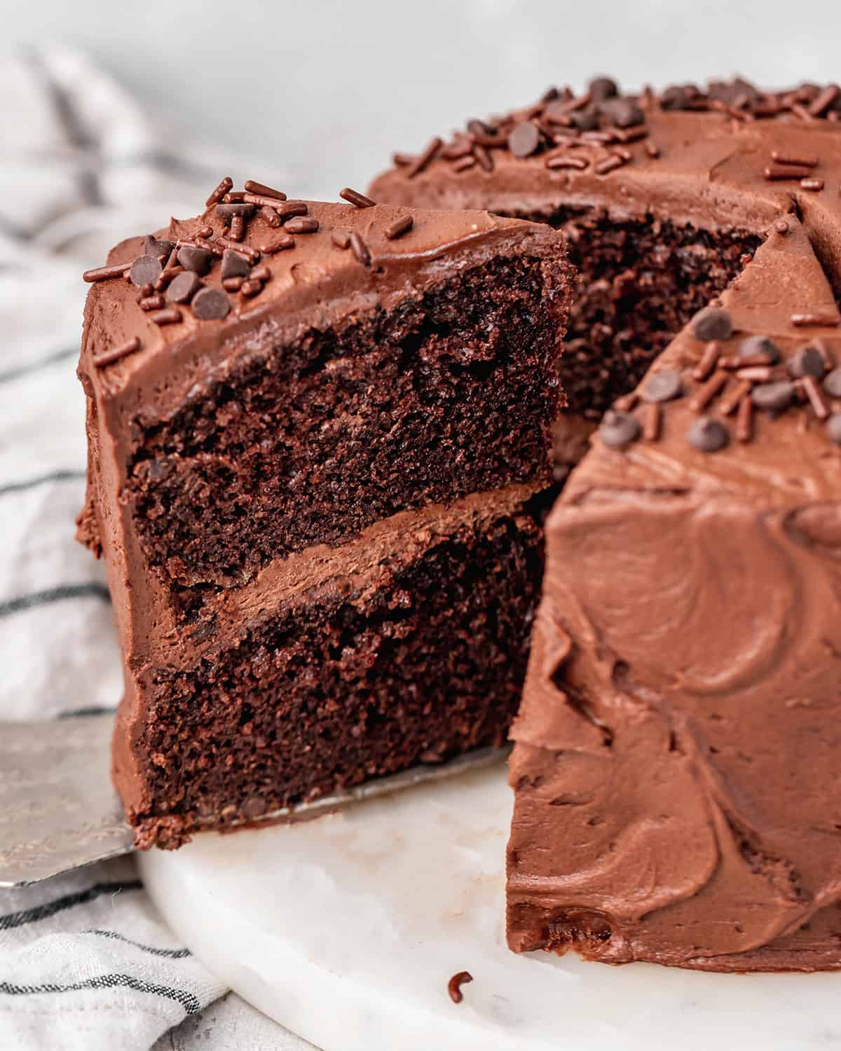 This is the best chocolate cake recipe for serious chocolate lovers. It's dense and moist with a perfect crumb! Topped with chocolate buttercream frosting it's the ultimate chocolate birthday cake. Check out the step-by-step photos and watch the video to learn how to make this easy chocolate cake recipe and ditch boxed cake mixes once and for all! a slice of Chocolate Cake being taken out of the whole cake with a cake server