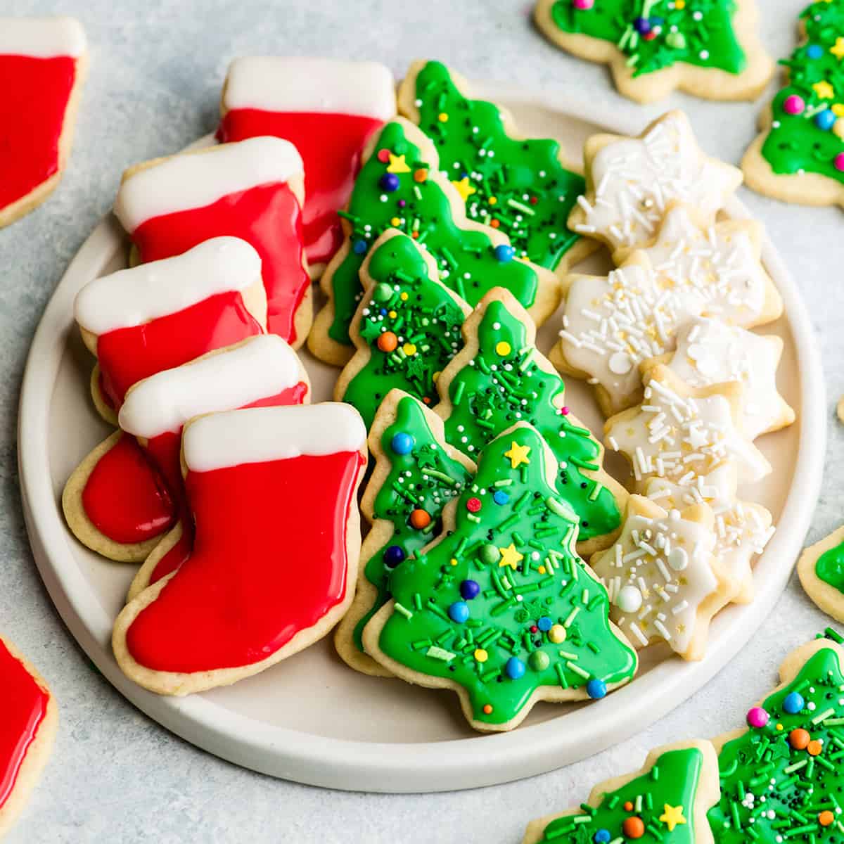 The Best Cut Out Sugar Cookie Recipe! They are soft, chewy and don't spread! Plus an easy sugar cookie frosting that tastes great and hardens with a beautiful shine that's perfect for decorating! Making these easy decorated sugar cookies has been our family's holiday baking tradition for 30 years! #sugarcookies #cutoutsugarcookies #christmascookies #christmas #baking #cutoutcookies #sugarcookiefrosting #recipe overhead view of a white, round plate with three rows of sugar cookies on it. The left row is five red and white stockings, the middle row is six green Christmas trees with sprinkles and the left row is seven small white stars with sprinkles.