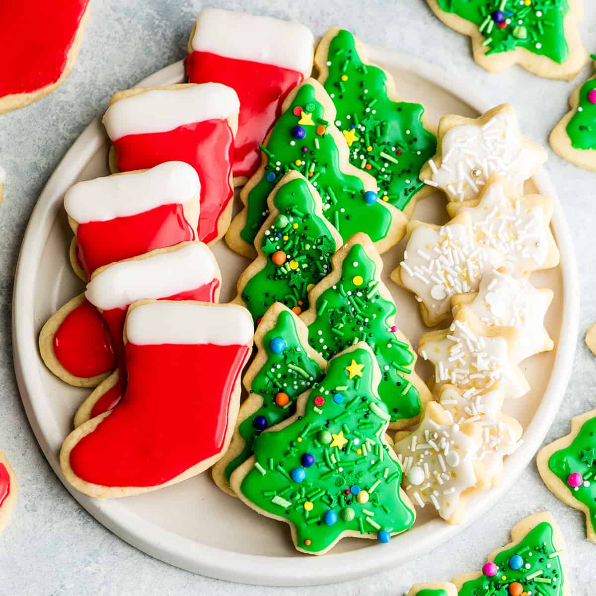 The Best Cut Out Sugar Cookie Recipe! They are soft, chewy and don't spread! Plus an easy sugar cookie frosting that tastes great and hardens with a beautiful shine that's perfect for decorating! Making these easy decorated sugar cookies has been our family's holiday baking tradition for 30 years! #sugarcookies #cutoutsugarcookies #christmascookies #christmas #baking #cutoutcookies #sugarcookiefrosting #recipe overhead view of a white, round plate with three rows of sugar cookies on it. The left row is five red and white stockings, the middle row is six green Christmas trees with sprinkles and the left row is seven small white stars with sprinkles.