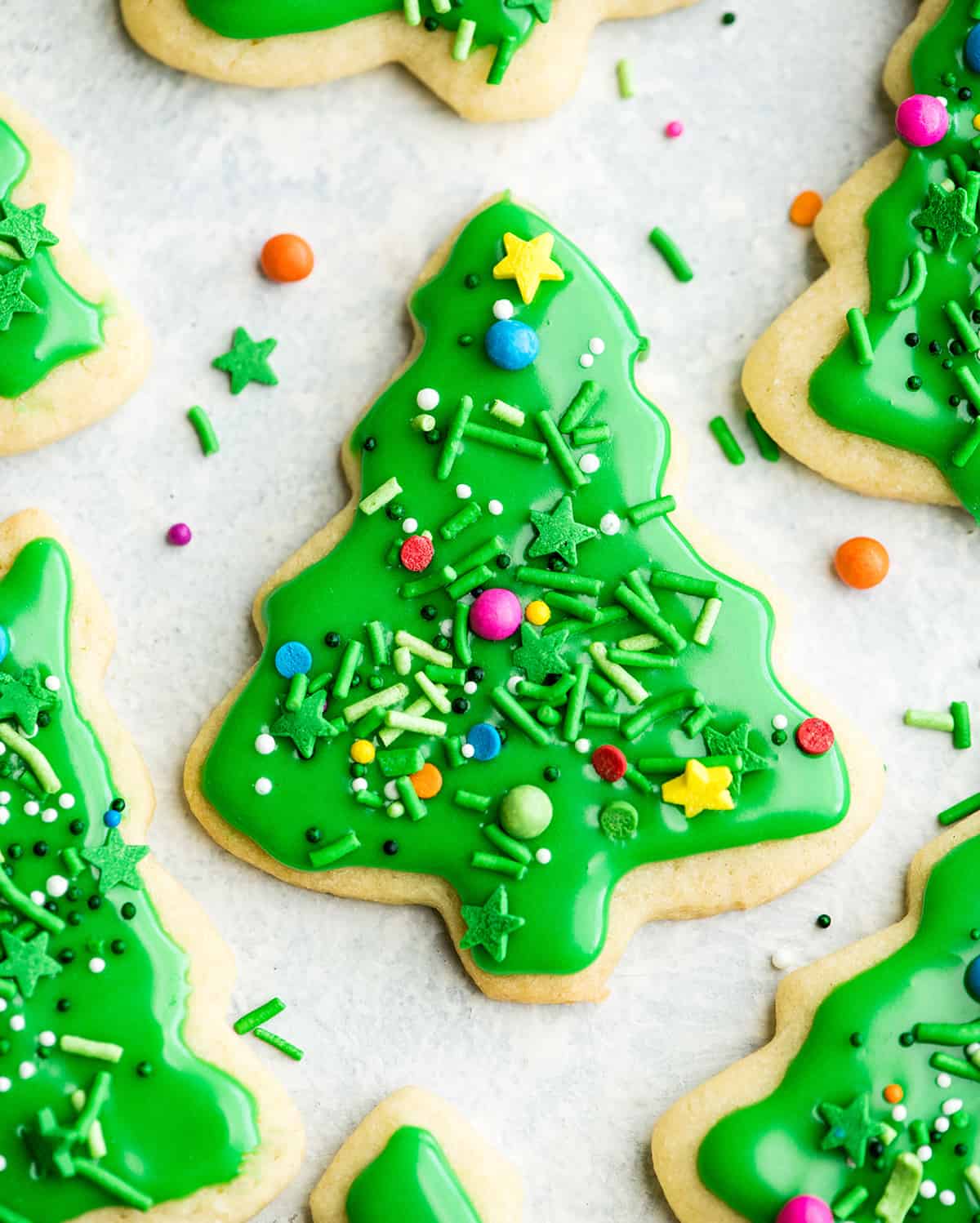 The Best Cut Out Sugar Cookie Recipe! They are soft, chewy and don't spread! Plus an easy sugar cookie frosting that tastes great and hardens with a beautiful shine that's perfect for decorating! Making these easy decorated sugar cookies has been our family's holiday baking tradition for 30 years! #sugarcookies #cutoutsugarcookies #christmascookies #christmas #baking #cutoutcookies #sugarcookiefrosting #recipe overhead view of a green Christmas tree sugar cookie with sprinkles