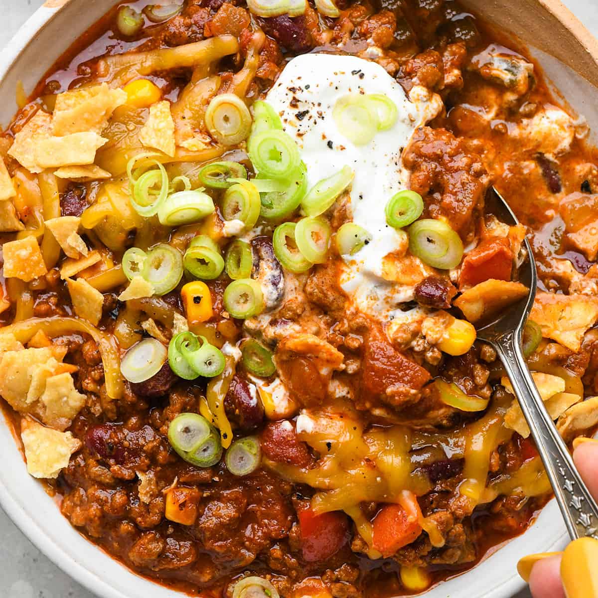a spoon taking a scoop of beef chili topped with sour cream, green onions, cheese and crushed chips.