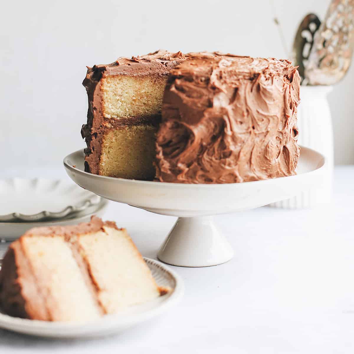 A Yellow Cake with chocolate frosting on a cake plate with a slice cut out on a plate in front of it
