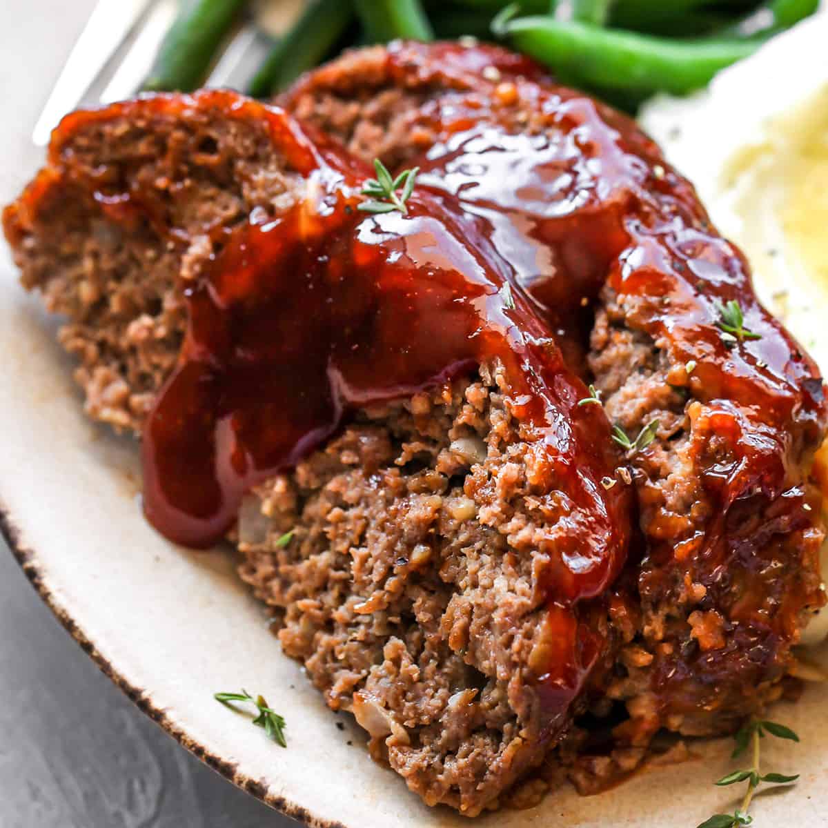 two slices of meatloaf on a plate with extra glaze, mashed potatoes and green beans