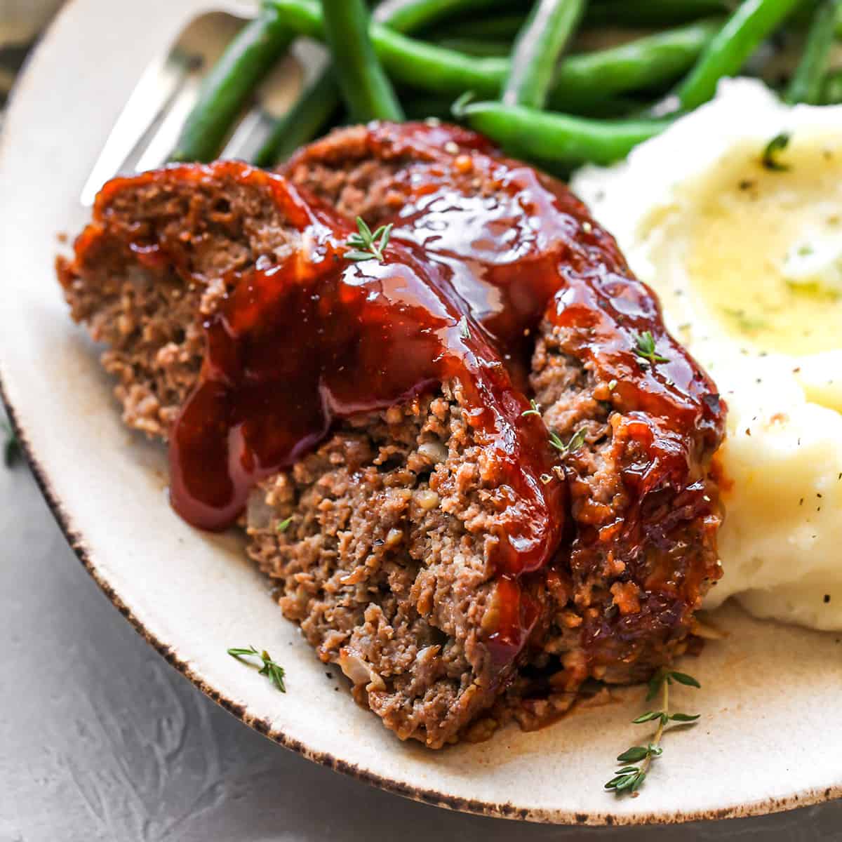 This is the best meatloaf recipe ever! It’s tender, moist and flavorful with the most delicious glaze. This easy meatloaf recipe is a classic that your whole family will love! Watch the video and follow our step-by-step instructions to learn how to make meatloaf! front view of two slices of the Best Meatloaf on a plate with beans and mashed potatoes