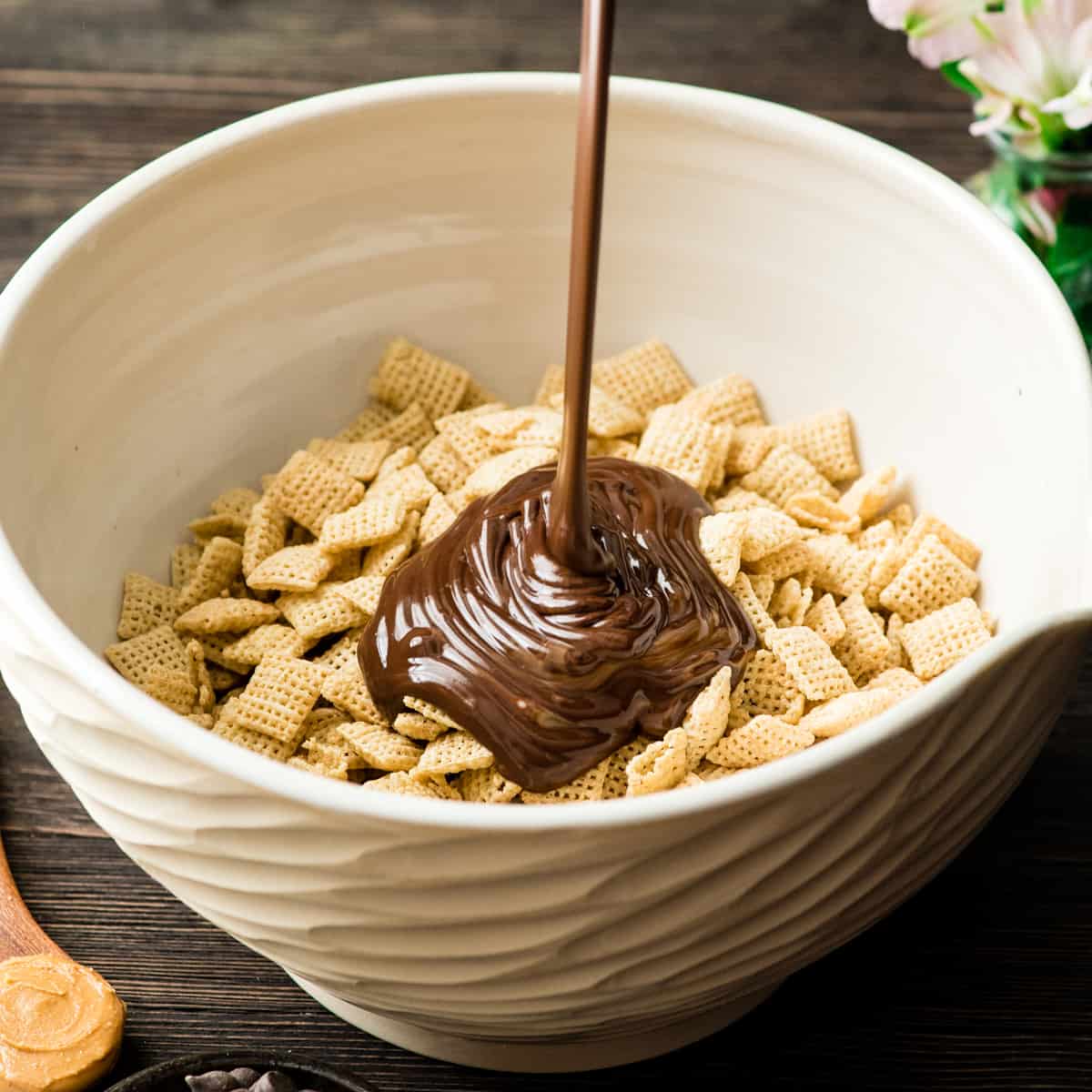 The Best Puppy Chow Recipe (AKA: Muddy Buddies) EVER! Only 4 ingredients (no butter) and a few minutes results in an irresistible dessert loaded with chocolate and peanut butter! The perfect sweet treat to feed a crowd! front view showing how to make puppy chow - pouring melted chocolate mixture onto cereal in a bowl