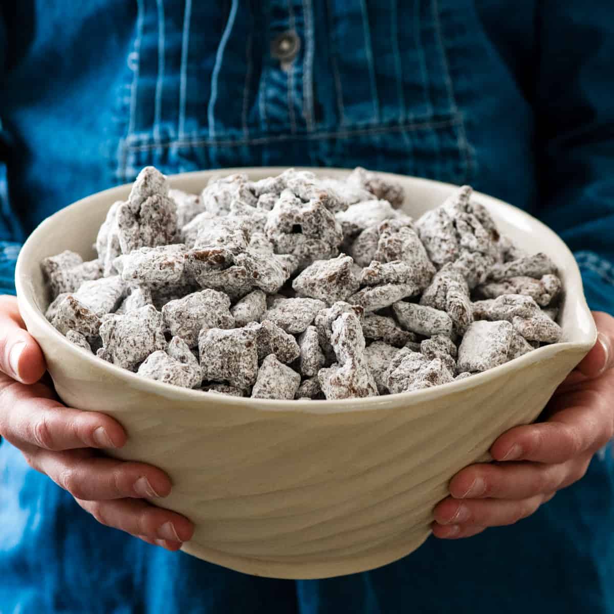 The Best Puppy Chow Recipe (AKA: Muddy Buddies) EVER! Only 4 ingredients (no butter) and a few minutes results in an irresistible dessert loaded with chocolate and peanut butter! The perfect sweet treat to feed a crowd! front view of hands holding a bowl of puppy chow (muddy buddies)