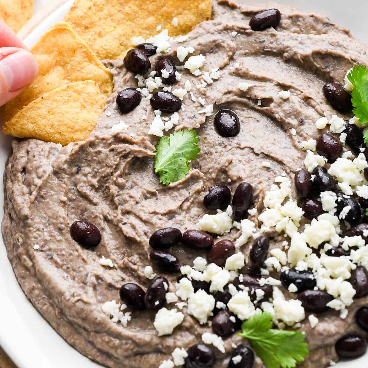 a hand dipping a chip into a bowl of Black Bean Dip garnished with cilantro. black beans, and cheese