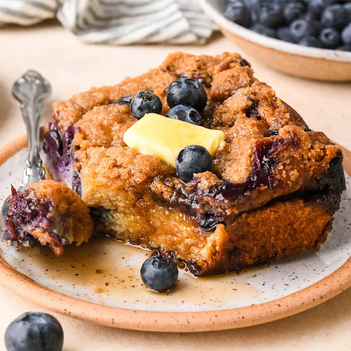 a piece of Blueberry French Toast Casserole on a plate with butter, syrup and a fork with a bite on it