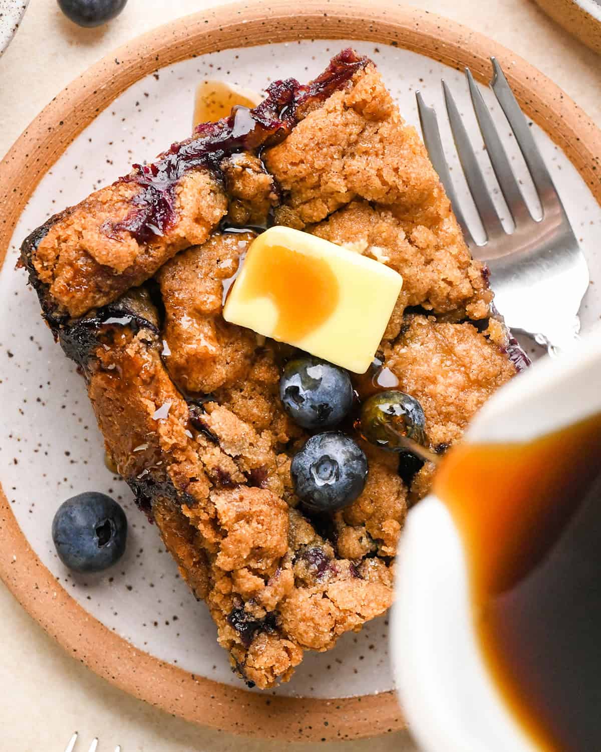 syrup being poured on a piece of Blueberry French Toast Casserole with butter and blueberries