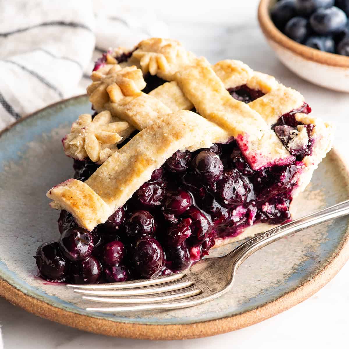 This is the best homemade Blueberry Pie Recipe you'll ever make! It's made from scratch with a fresh blueberry pie filling and NO cornstarch, and includes step-by-step instructions and photos! front view of a slice of blueberry pie on a plate