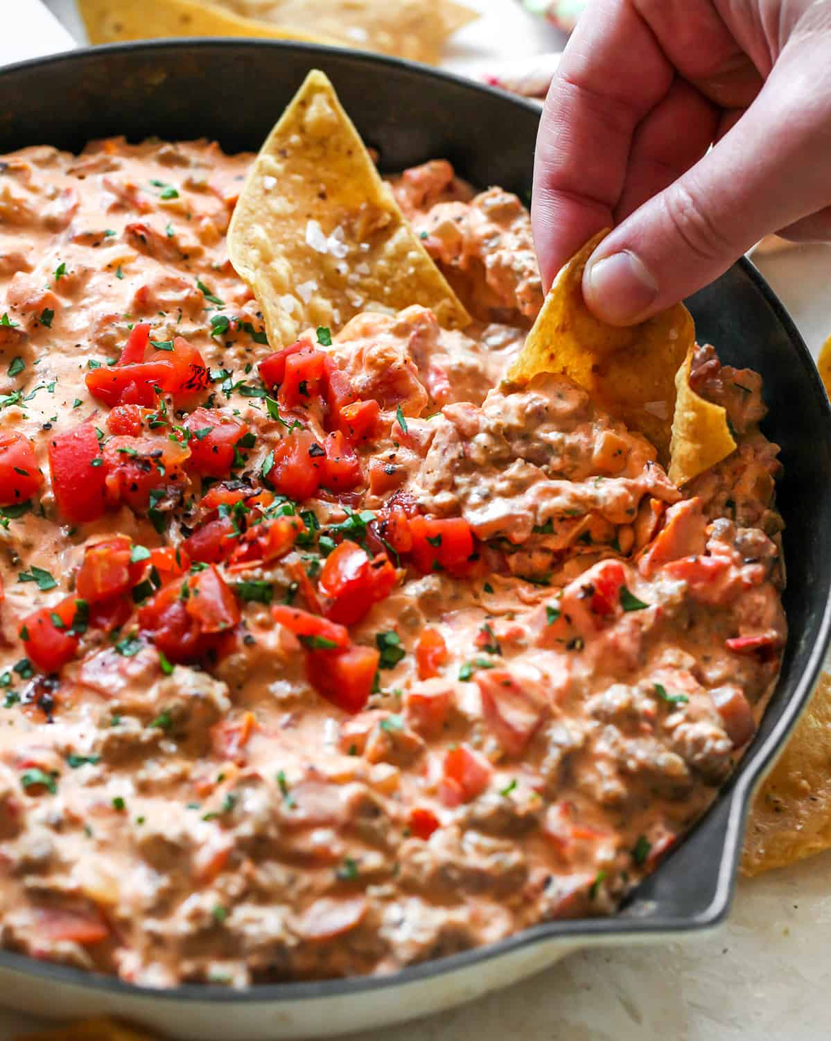 This cream cheese sausage dip is an easy appetizer made with 8 ingredients, in one skillet in 20 minutes. You can easily adjust the spice to make it mild or hot! Serve it with chips, crackers, or veggies for a delicious, low-carb, warm appetizer that will be the hit of any party. front view of a chip being dipped into a skillet of Cream Cheese Sausage Dip