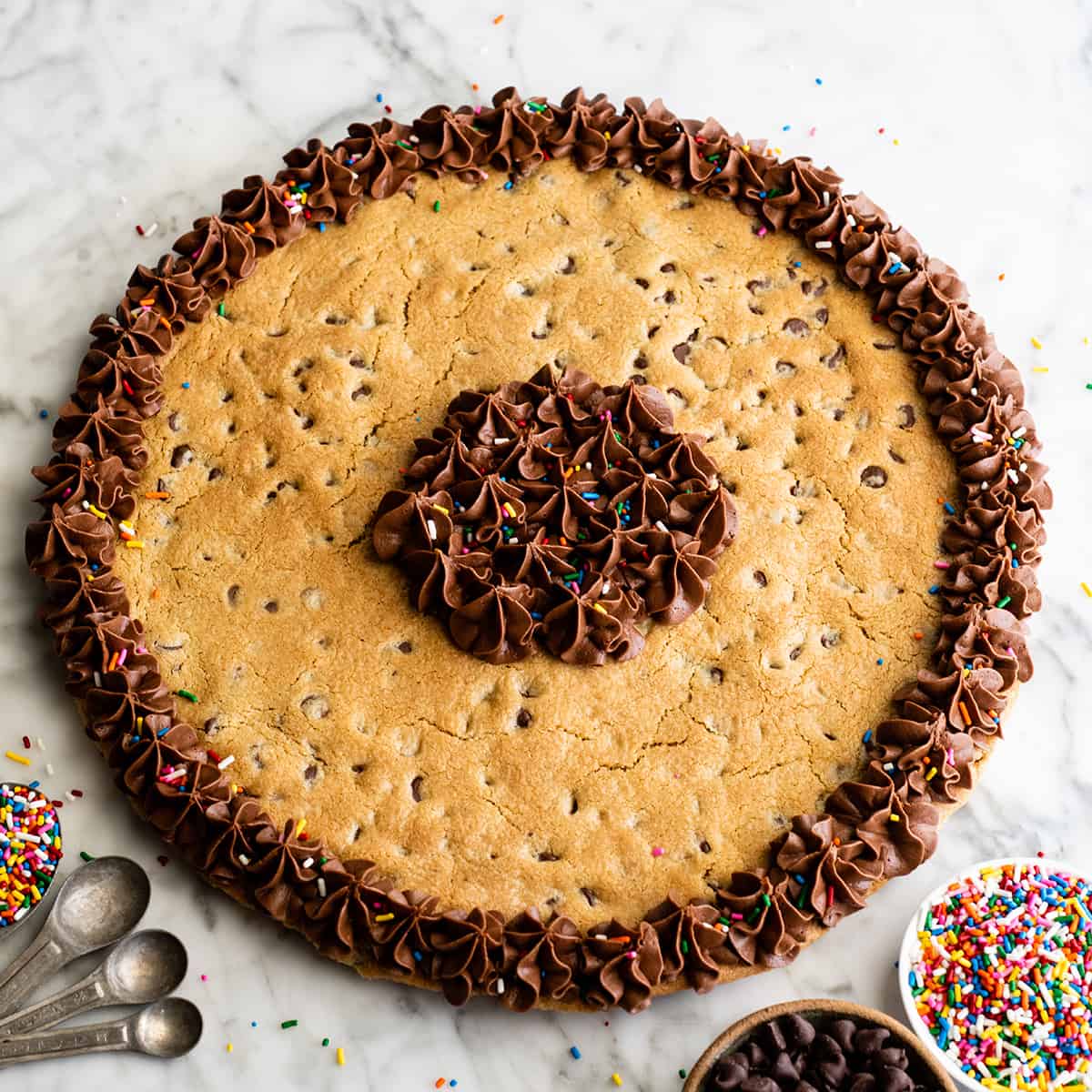 This chocolate chip cookie cake recipe is soft, chewy, slightly doughy and decorated with the best chocolate buttercream frosting. It's easy to make, large enough to feed a crowd, and even better than store-bought! photo of a Chocolate Chip Cookie Cake decorated with chocolate frosting
