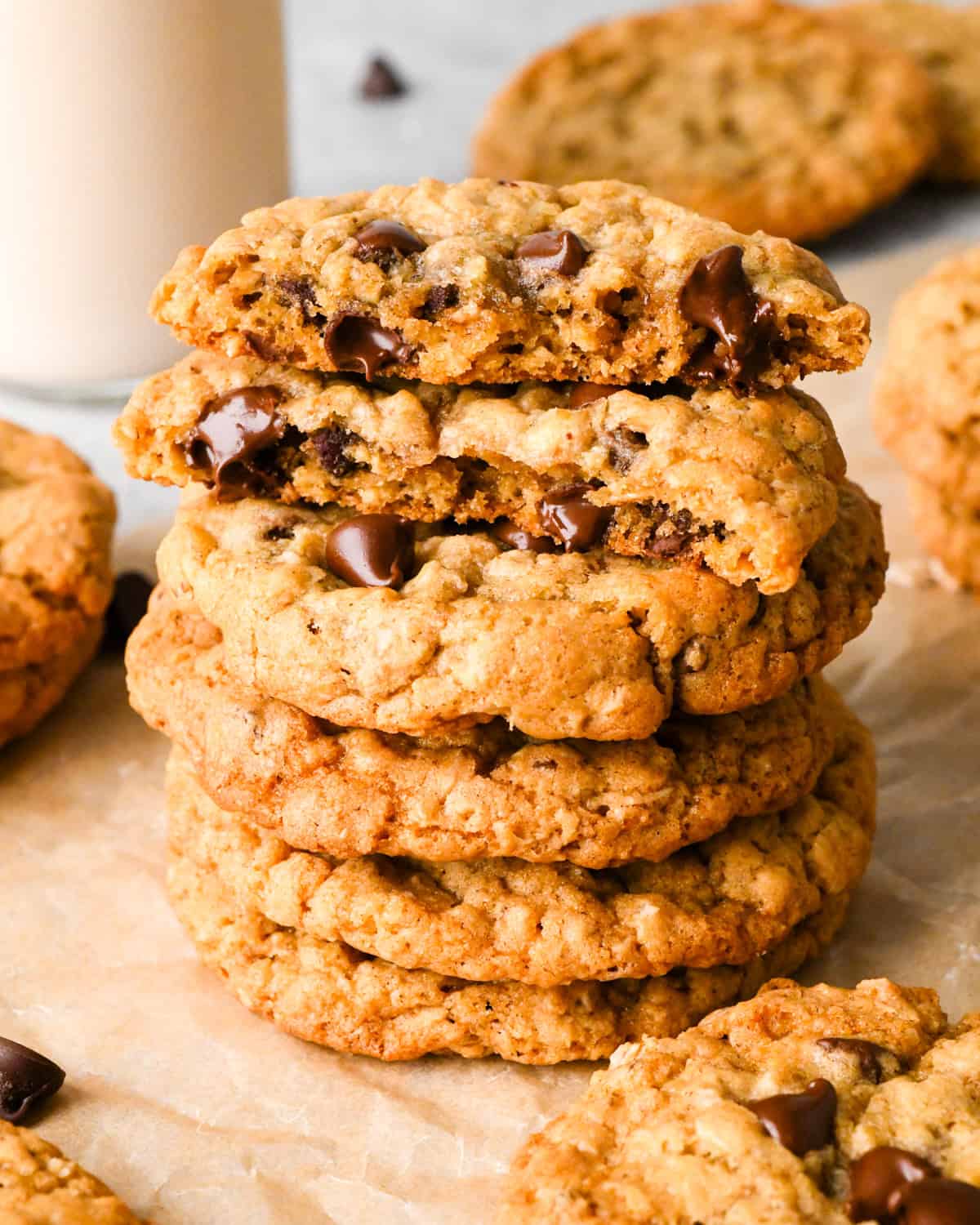 a stack of 4 Chocolate Chip Oatmeal Cookies on parchment paper, the top one is cut in half and the halves are stacked on each other. Surrounded by other cookies and chocolate chips