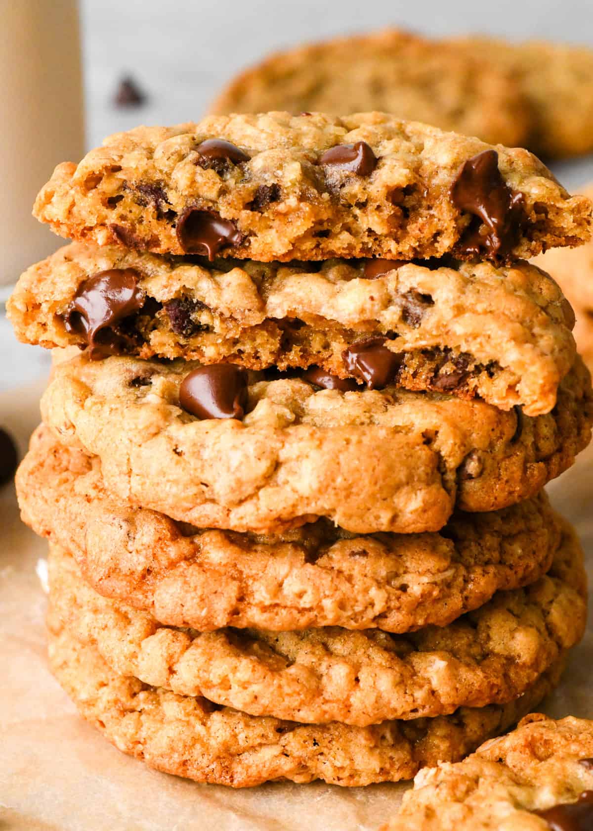 a stack of 5 Oatmeal Chocolate Chip Cookies on parchment paper, the top one is cut in half and the halves are stacked on each other. Surrounded by other cookies and chocolate chips