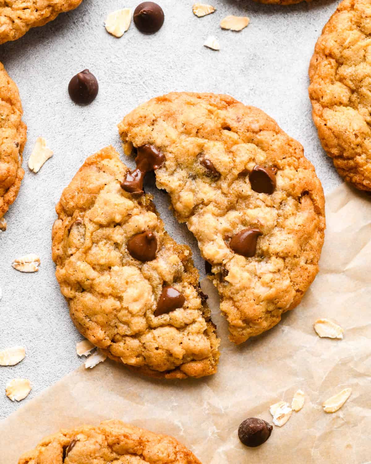 4 Oatmeal Chocolate Chip Cookies on parchment paper, one cut in half, surrounded by raw oats and chocolate chips.