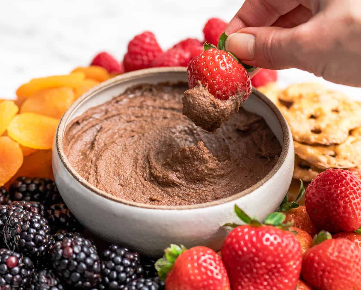 Healthy Chocolate Hummus Recipe is easy to make and ready in 5 minutes! It's a nutritious dip, appetizer, dessert, snack or spread that everyone in my family, including my kids, loves. This dessert hummus recipe is gluten-free, dairy-free, has no refined sugar & it's vegan! #hummus #chocolatehummus #vitamix #desserthummus #vegan Front view of a hand dipping a strawberry into a bowl of Chocolate Hummus
