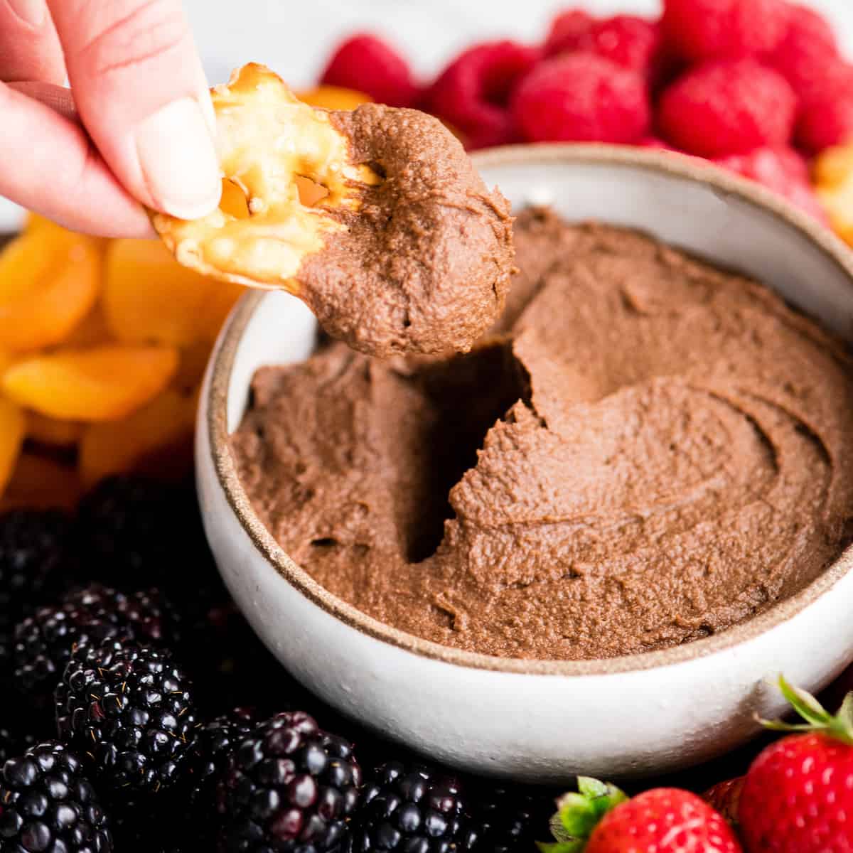 Healthy Chocolate Hummus Recipe is easy to make and ready in 5 minutes! It's a nutritious dip, appetizer, dessert, snack or spread that everyone in my family, including my kids, loves. This dessert hummus recipe is gluten-free, dairy-free, has no refined sugar & it's vegan! #hummus #chocolatehummus #vitamix #desserthummus #vegan Front view of a hand dipping a pretzel chip into a bowl of Chocolate Hummus