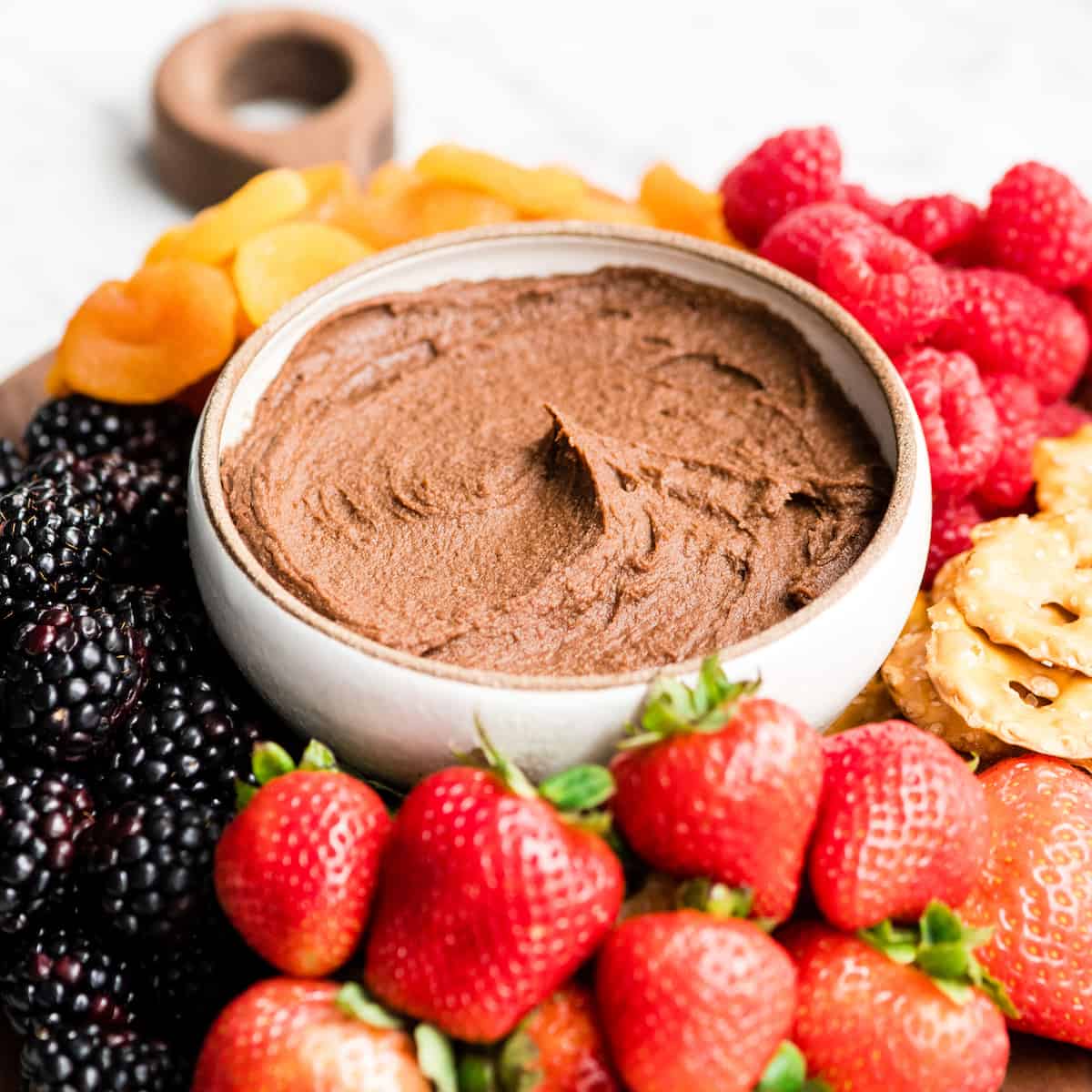 Healthy Chocolate Hummus Recipe is easy to make and ready in 5 minutes! It's a nutritious dip, appetizer, dessert, snack or spread that everyone in my family, including my kids, loves. This dessert hummus recipe is gluten-free, dairy-free, has no refined sugar & it's vegan! #hummus #chocolatehummus #vitamix #desserthummus #vegan Front view of a bowl of chocolate hummus surrounded by fruits, dried fruits and pretzels