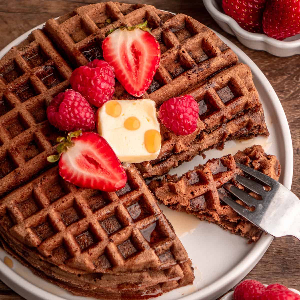 Chocolate Waffles on a plate with butter, berries and syrup and a fork taking a bite
