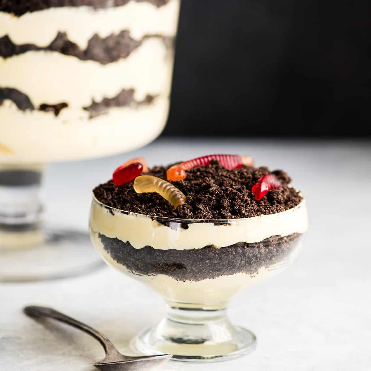 This Easy Dirt Cake Recipe (Oreo Dirt Pudding) is one of our favorite no-bake desserts! Crushed Oreos are layered between a french vanilla pudding mixture for a kid-friendly, make-ahead dessert recipe perfect for any potluck or BBQ! Add gummy worms for a whimsical touch! #dirtcake #dirtpudding #oreos #oreopudding #nobake #dessert #potluck #bbq #summer Front view of a small glass dish of Oreo Dirt Cake with two layers of pudding and two layers of crushed oreos with gummy worms on top. A large glass trifle dish of Oreo Dirt Cake is in the background and a silver spoon is in the foreground
