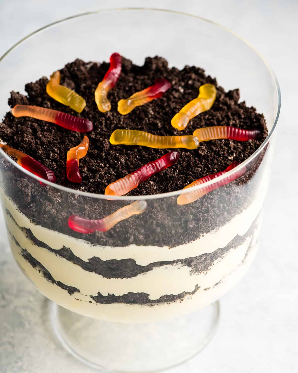 This Easy Dirt Cake Recipe (Oreo Dirt Pudding) is one of our favorite no-bake desserts! Crushed Oreos are layered between a french vanilla pudding mixture for a kid-friendly, make-ahead dessert recipe perfect for any potluck or BBQ! Add gummy worms for a whimsical touch! #dirtcake #dirtpudding #oreos #oreopudding #nobake #dessert #potluck #bbq #summer Front view of this Oreo Dirt Cake Recipe in a glass trifle dish with three layers of pudding and three layers of crushed Oreos with gummy worms on top