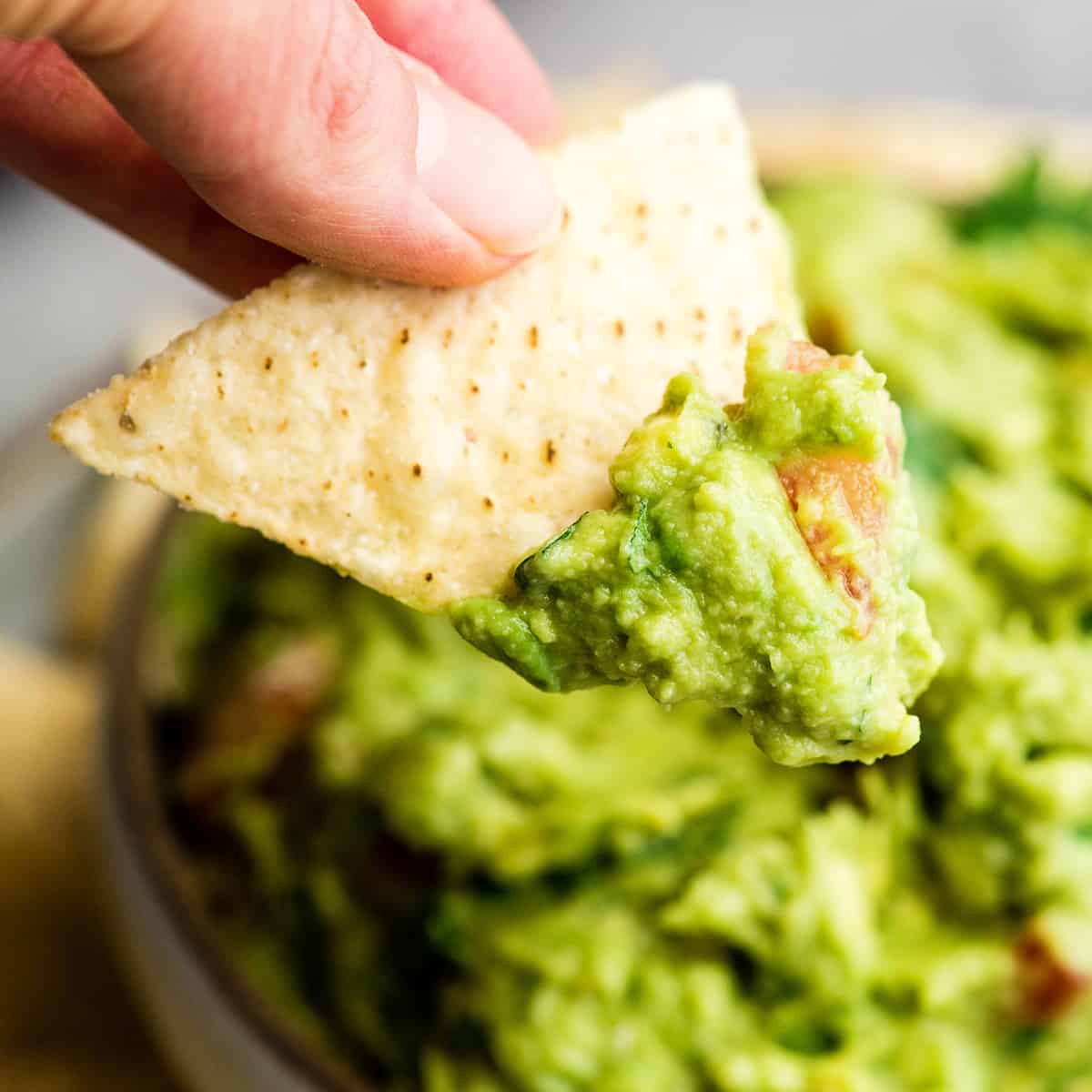 This quick & easy guacamole recipe is made with a handful of healthy ingredients and is ready in 5 minutes! This simple guacamole dip is our favorite appetizer and we always make it on taco nights! Watch the video to learn how to make this guacamole recipe! #guacamole #guacamolerecipe #tacos #appetizer #dip up close front view of a hand holding a chip with guacamole on it