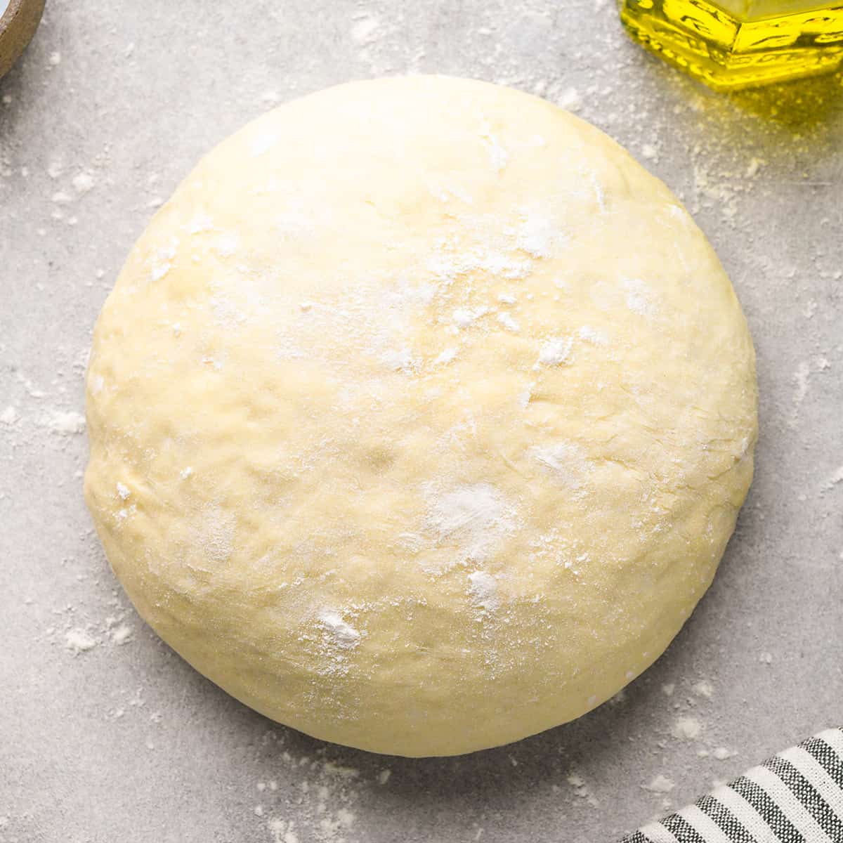 a ball of homemade pizza dough on a floured gray floured surface with olive oil and a striped towel nearby
