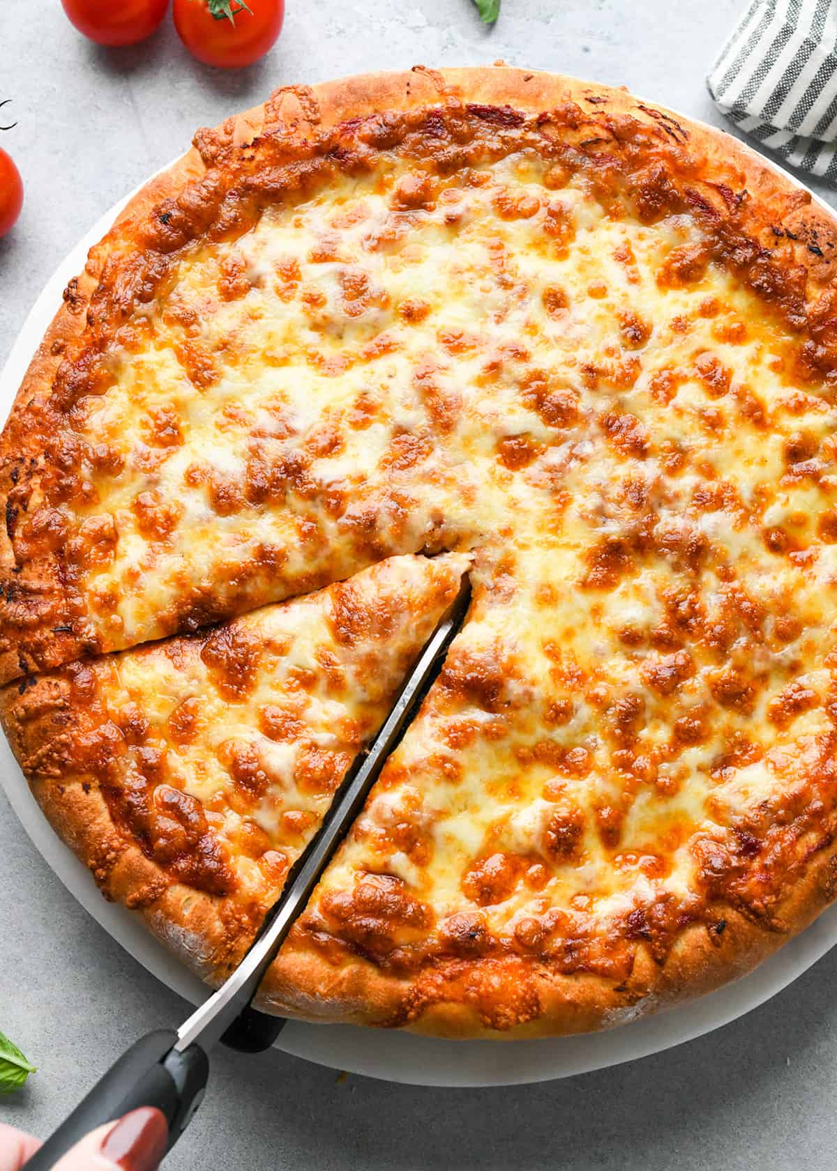 pizza made with homemade pizza dough being cut with pizza scissors with tomatoes, basil and a striped towel nearby