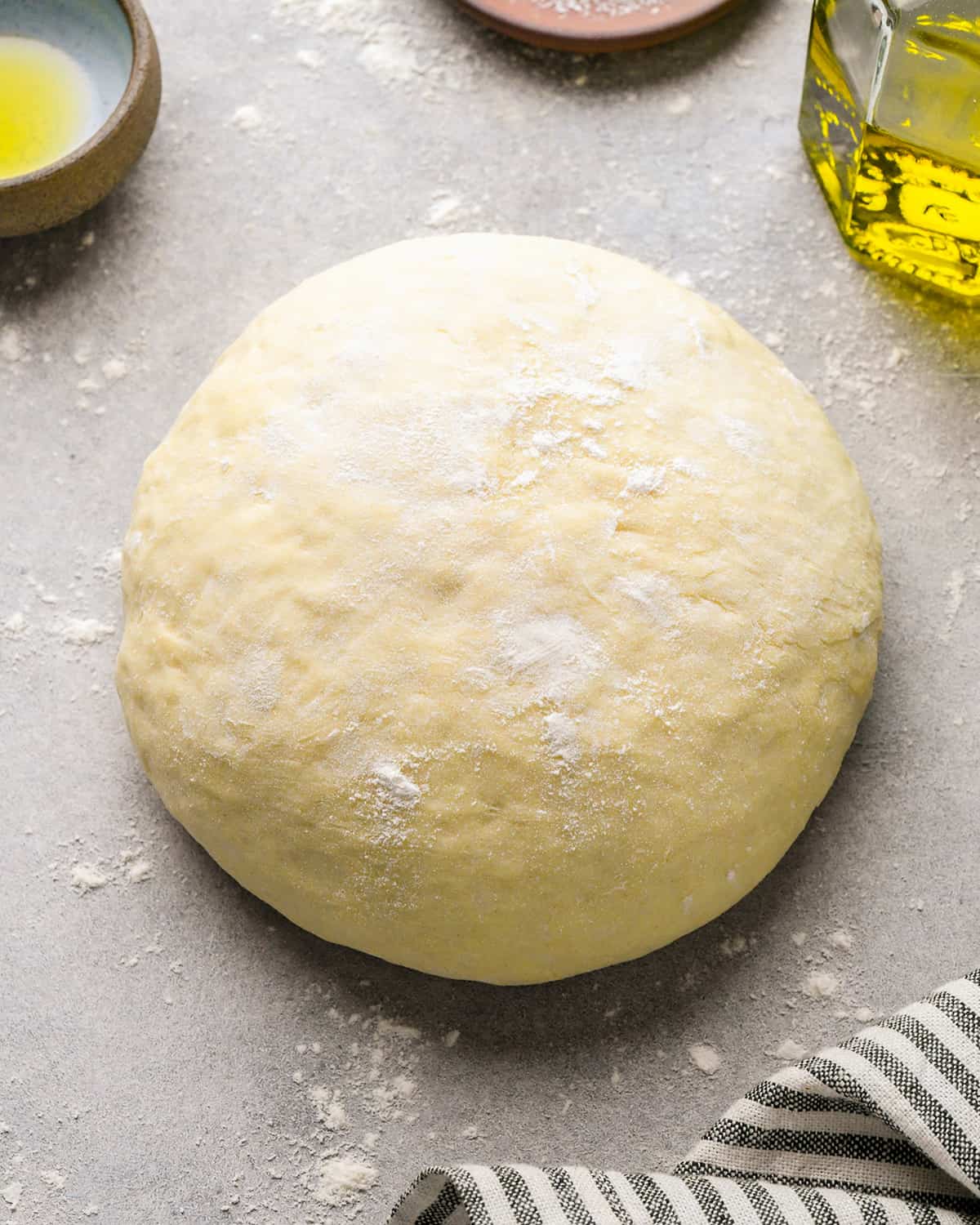 a ball of homemade pizza dough on a floured gray floured surface with olive oil and a striped towel nearby