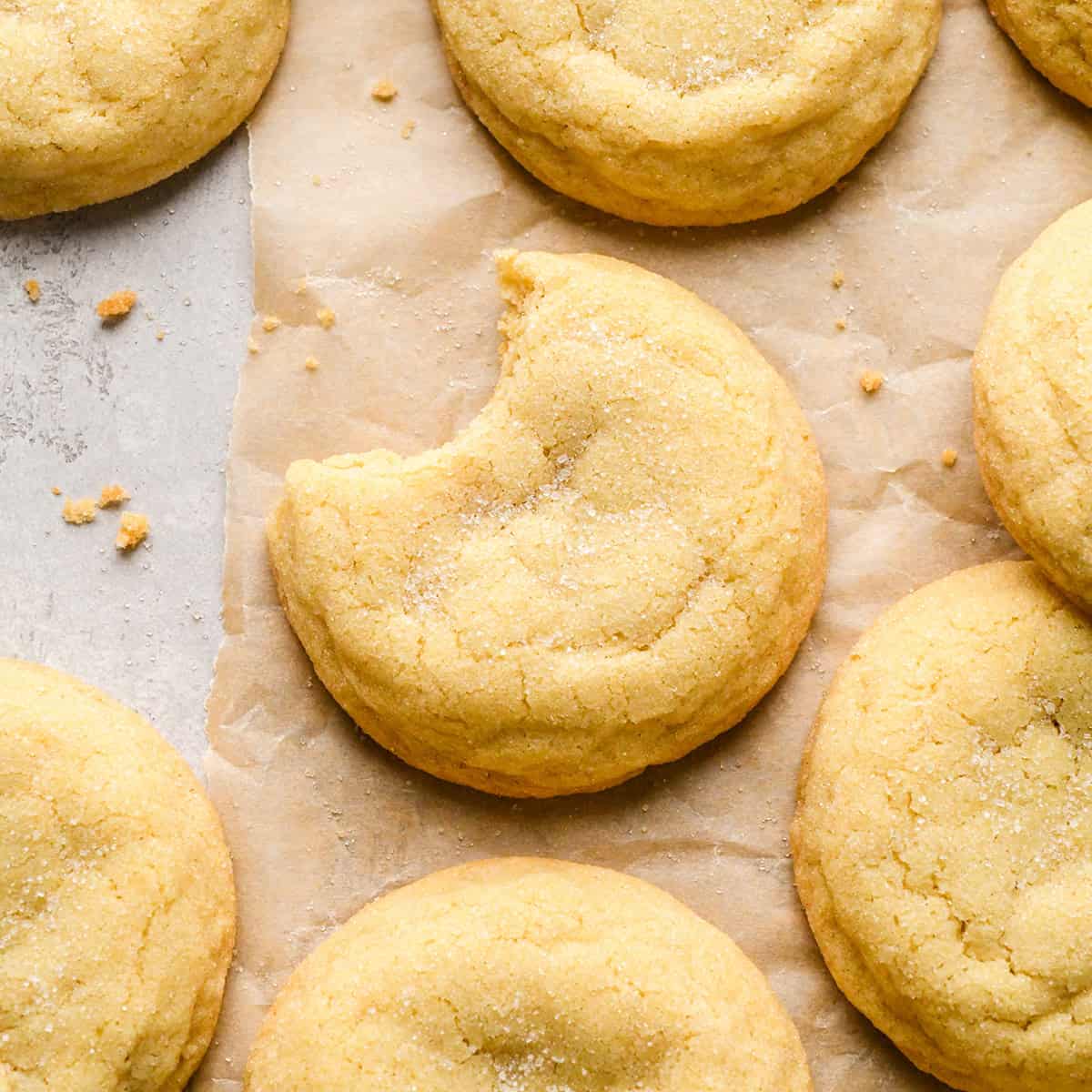 7 sugar cookies on parchment paper sprinkled with granulated sugar with crumbs around them. The middle cookie has a bite taken out of it