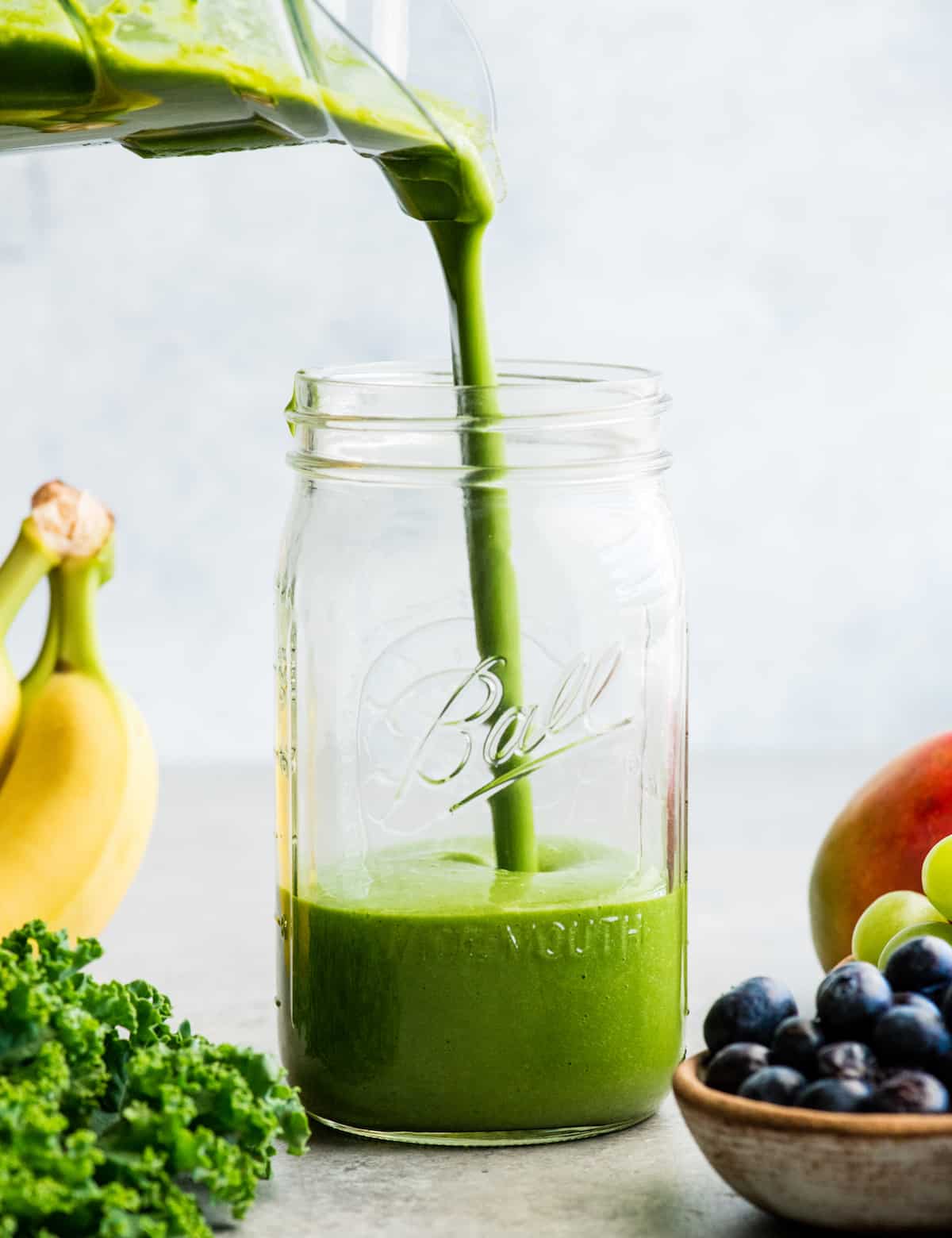 How to make the best green smoothie recipe! A simple green smoothie for beginners that is healthy, tastes amazing, and it's made with 4 ingredients in less than 5 minutes! Plus my tips on meal prepping smoothies! #greensmoothie #smoothie #smoothierecipe #greensmoothierecipe #kidfriendly front view of a green smoothie being poured into a large glass mason jar, surrounded with kale, blueberries, bananas, mangoes and green grapes