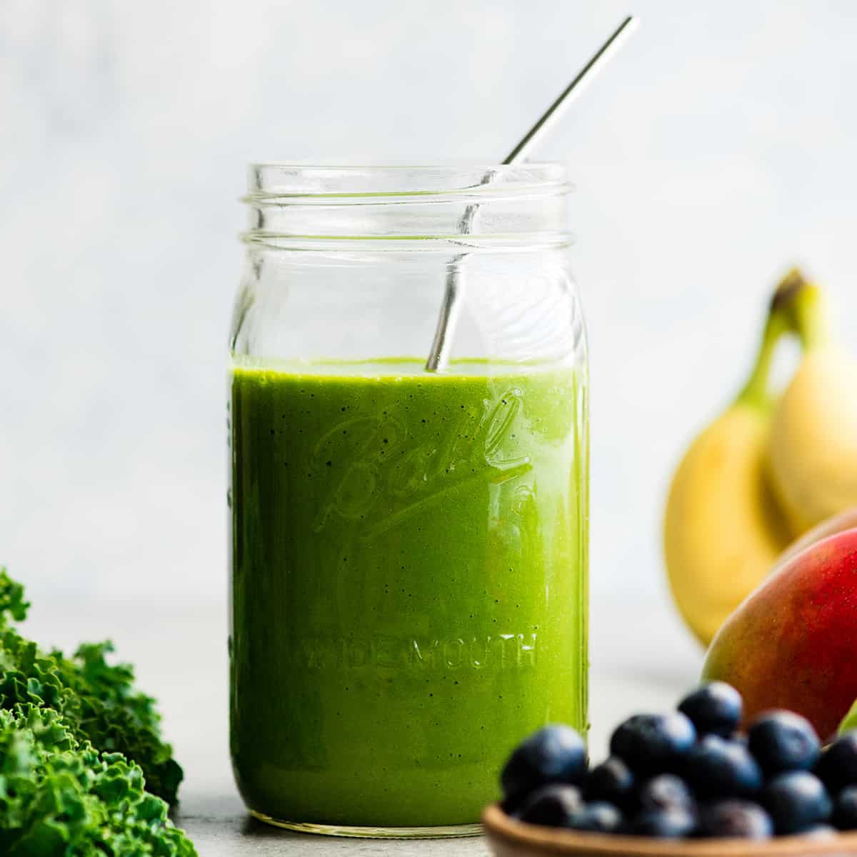 How to make the best green smoothie recipe! A simple green smoothie for beginners that is healthy, tastes amazing, and it's made with 4 ingredients in less than 5 minutes! Plus my tips on meal prepping smoothies! #greensmoothie #smoothie #smoothierecipe #greensmoothierecipe #kidfriendly front view of a green smoothie in a large glass mason jar with a metal straw, surrounded with kale, blueberries, bananas, mangoes and green grapes