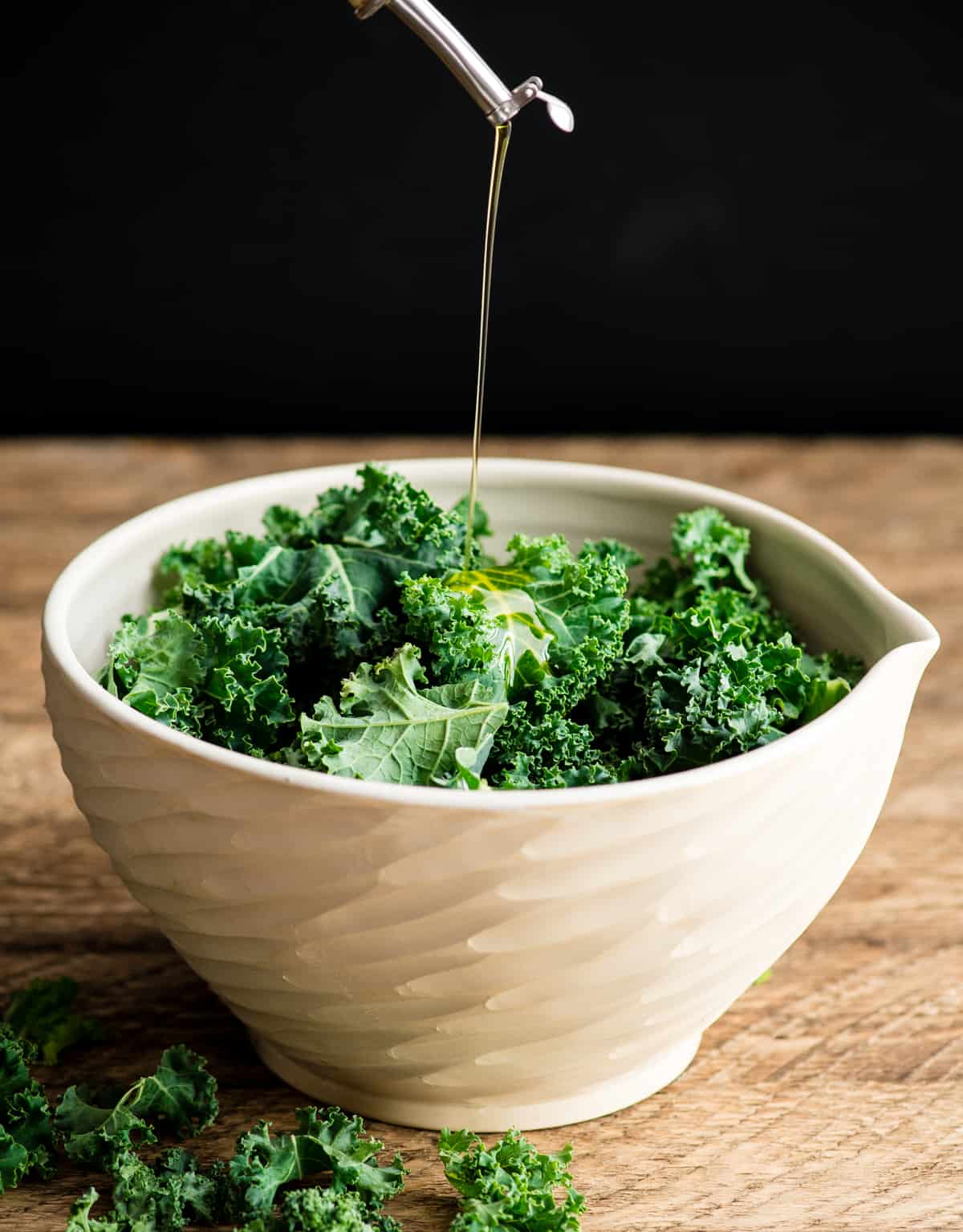 This is the best Baked Kale Chips recipe ever! Homemade crispy kale chips are a delicious, healthy snack made with only five ingredients. Roasting kale chips in the oven keeps them crispy for days! Front view of olive oil being poured into a bowl of chopped kale leaves making baked kale chips