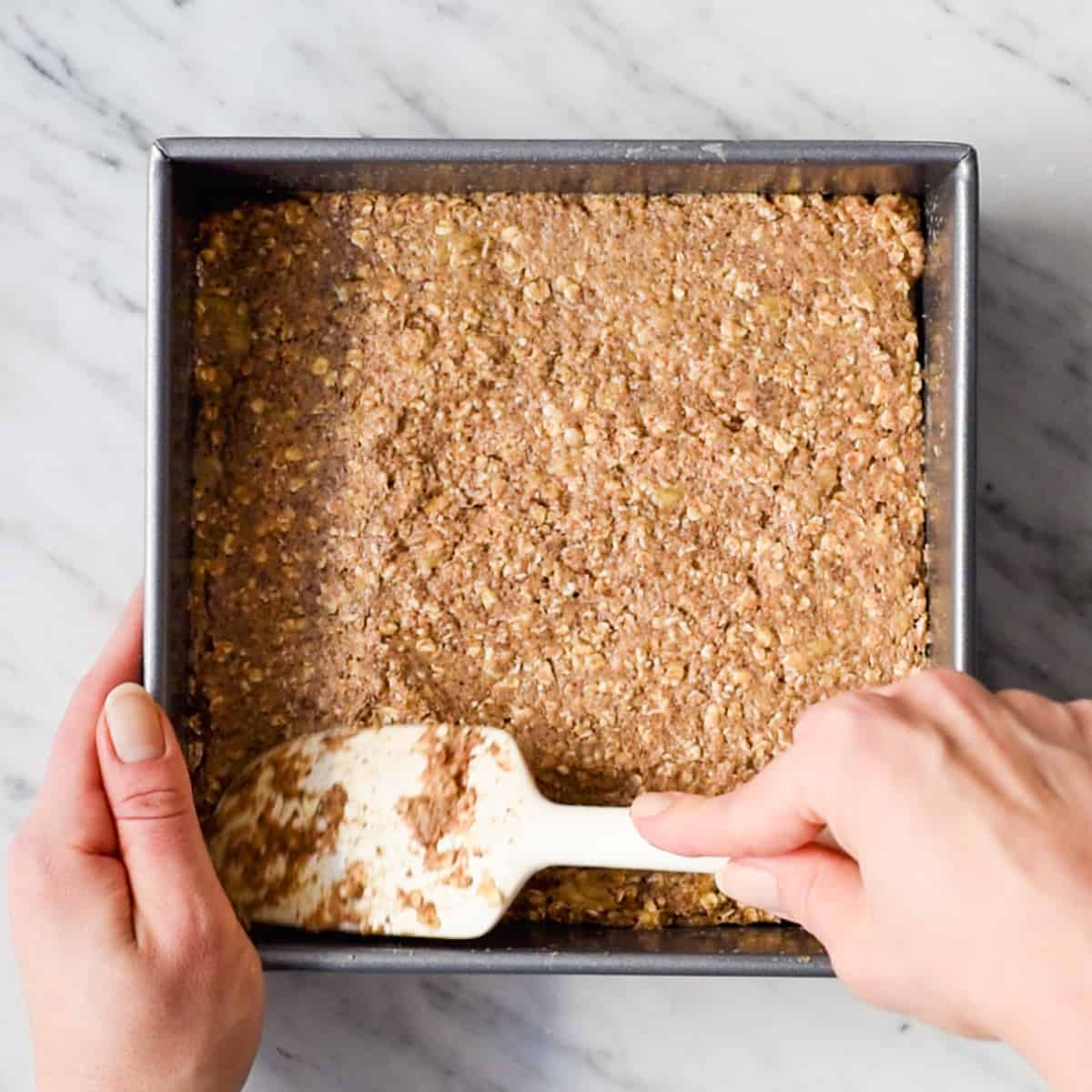 These are the BEST Healthy Breakfast Bars ever! I have eaten one every day for over a decade (seriously)! This easy recipe is made in one bowl in 30 minutes, and is loaded with fiber, protein & omega-3s to keep you full all morning long! Plus they’re gluten-free, dairy-free, refined sugar free and vegan! #breakfastbars #oatmeal #peanutbutter #glutenfree #dairyfree #vegan #healthy #refinedsugarfree overhead view of hands spreading the breakfast bar batter into a square pan with a white spatula before baking.