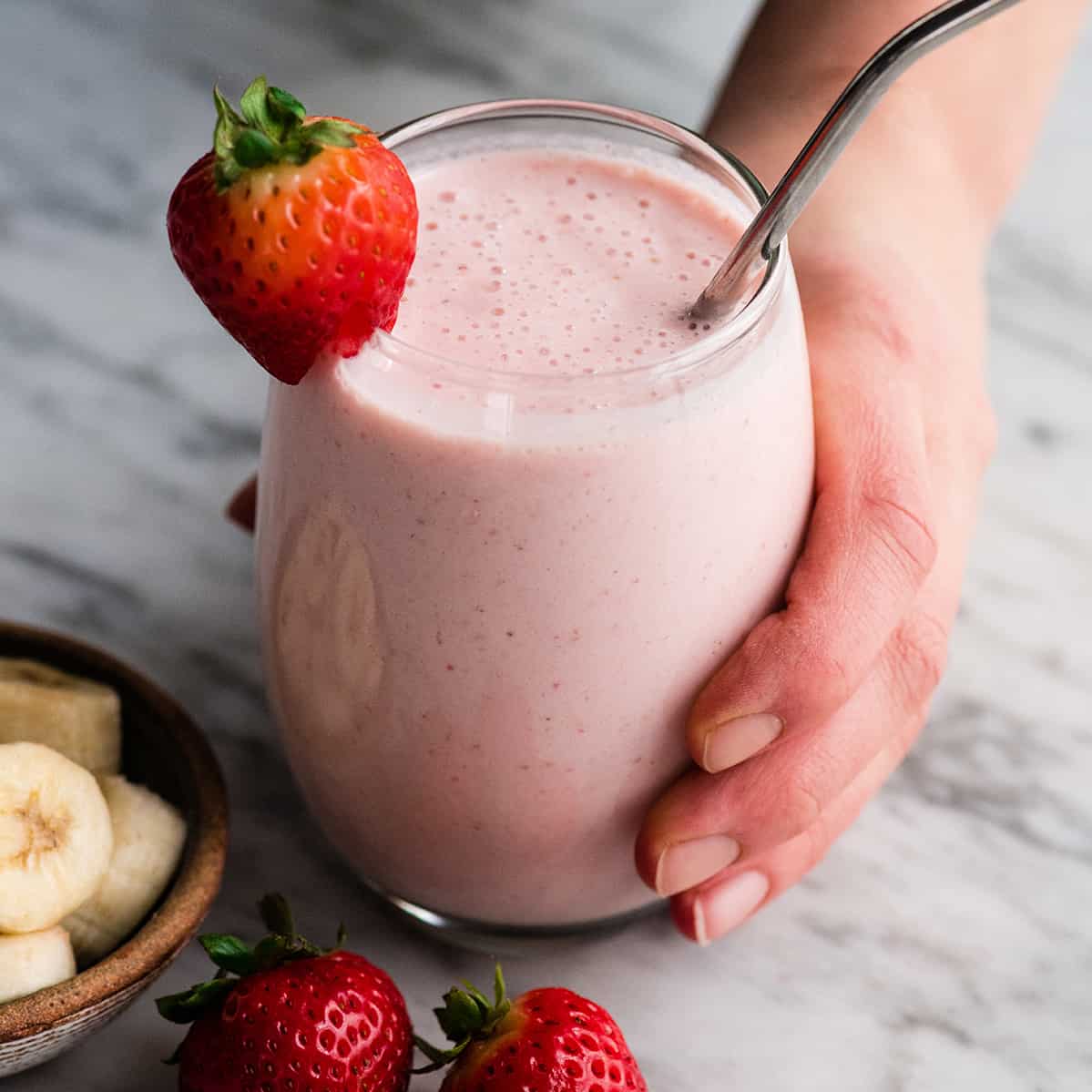 The BEST strawberry banana smoothie recipe ever! It's easy (ready in less than 5 minutes), healthy, and made with yogurt so it's full of protein! It's the perfect snack or breakfast! #smoothie #strawberrybananasmoothie #strawberry #banana #breakfast #snack #vitamix hand holding a glass filled with strawberry banana smoothie with a straw and strawberries.