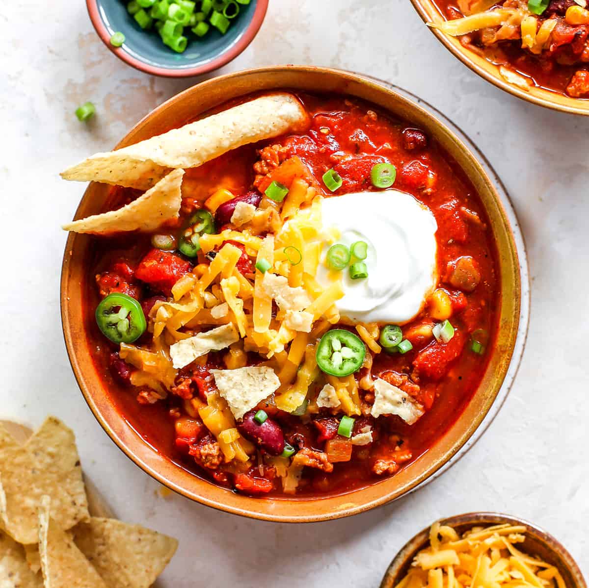 This Healthy Turkey Chili recipe is easy to make in 30 minutes on the stovetop. Turkey chili is loaded with protein and veggies and bursting with flavor thanks to a homemade spice mixture. A nutritious and cozy fall meal. overhead photo of a bowl of turkey chili with sour cream, cheese, chips and green onions