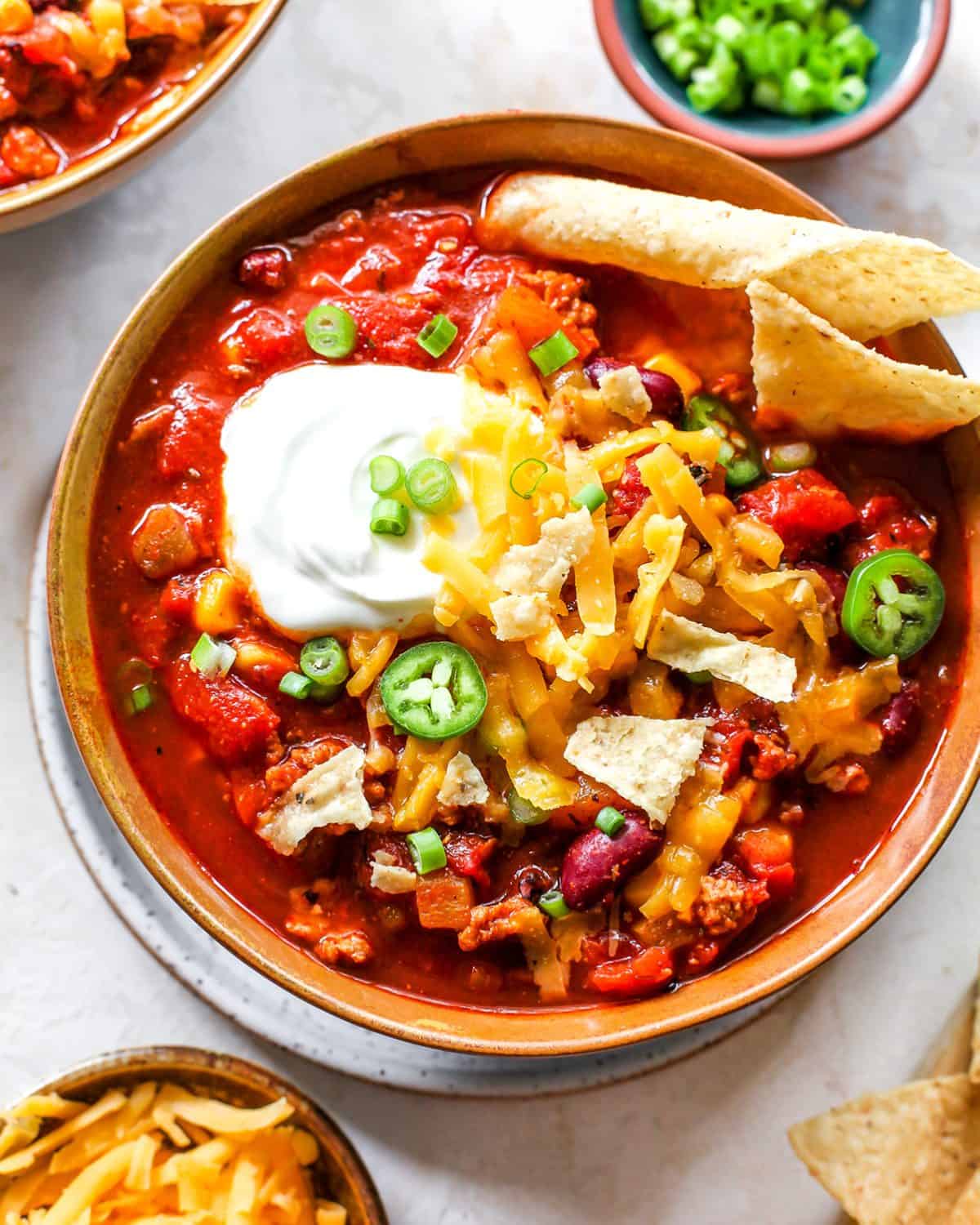 This Healthy Turkey Chili recipe is easy to make in 30 minutes on the stovetop. Turkey chili is loaded with protein and veggies and bursting with flavor thanks to a homemade spice mixture. A nutritious and cozy fall meal. overhead view of a bowl of turkey chili with sour cream, cheese, chips and green onions