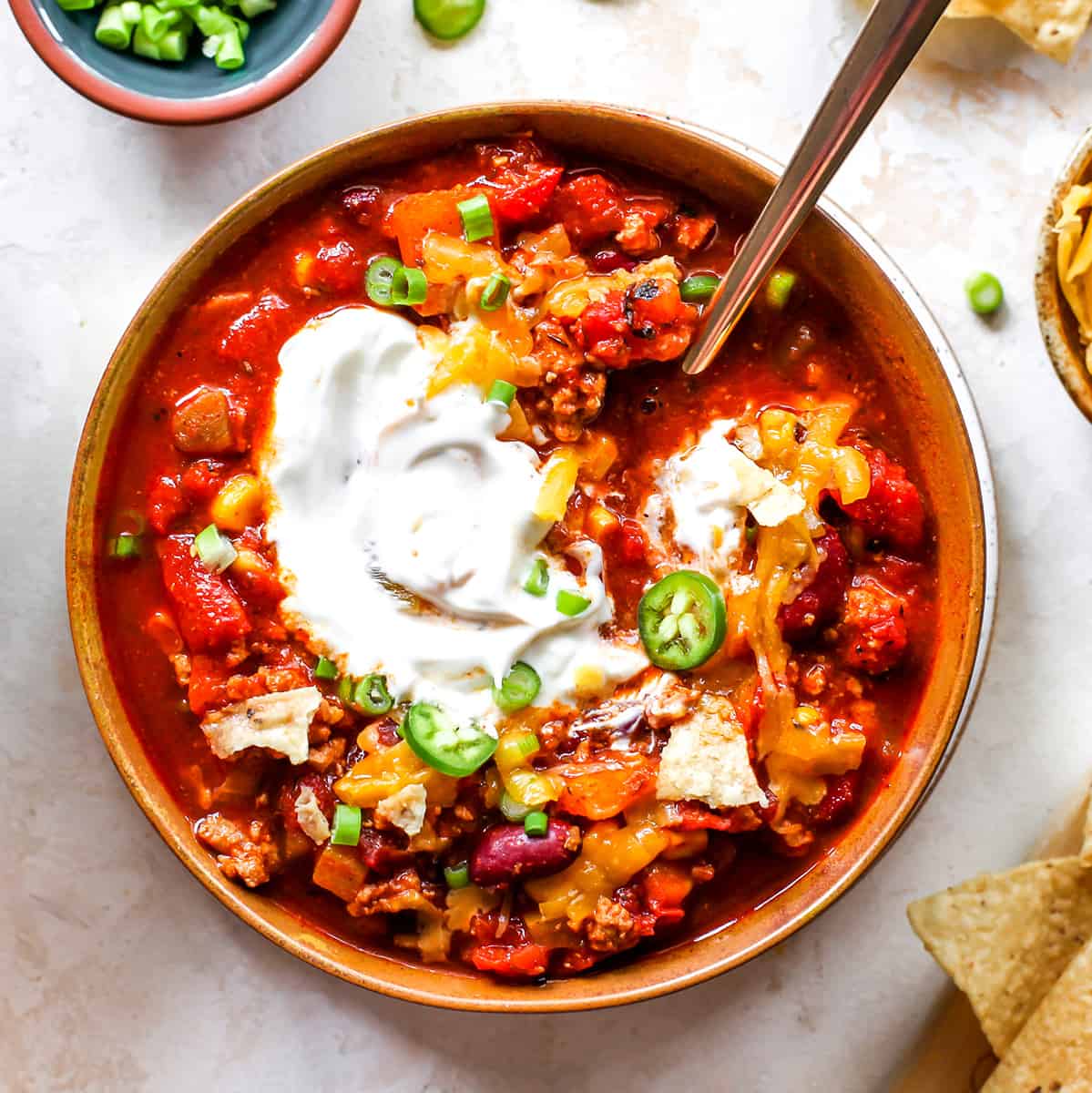 This Healthy Turkey Chili recipe is easy to make in 30 minutes on the stovetop. Turkey chili is loaded with protein and veggies and bursting with flavor thanks to a homemade spice mixture. A nutritious and cozy fall meal. Overhead photo of a bowl of turkey chili with sour cream, cheese and green onions