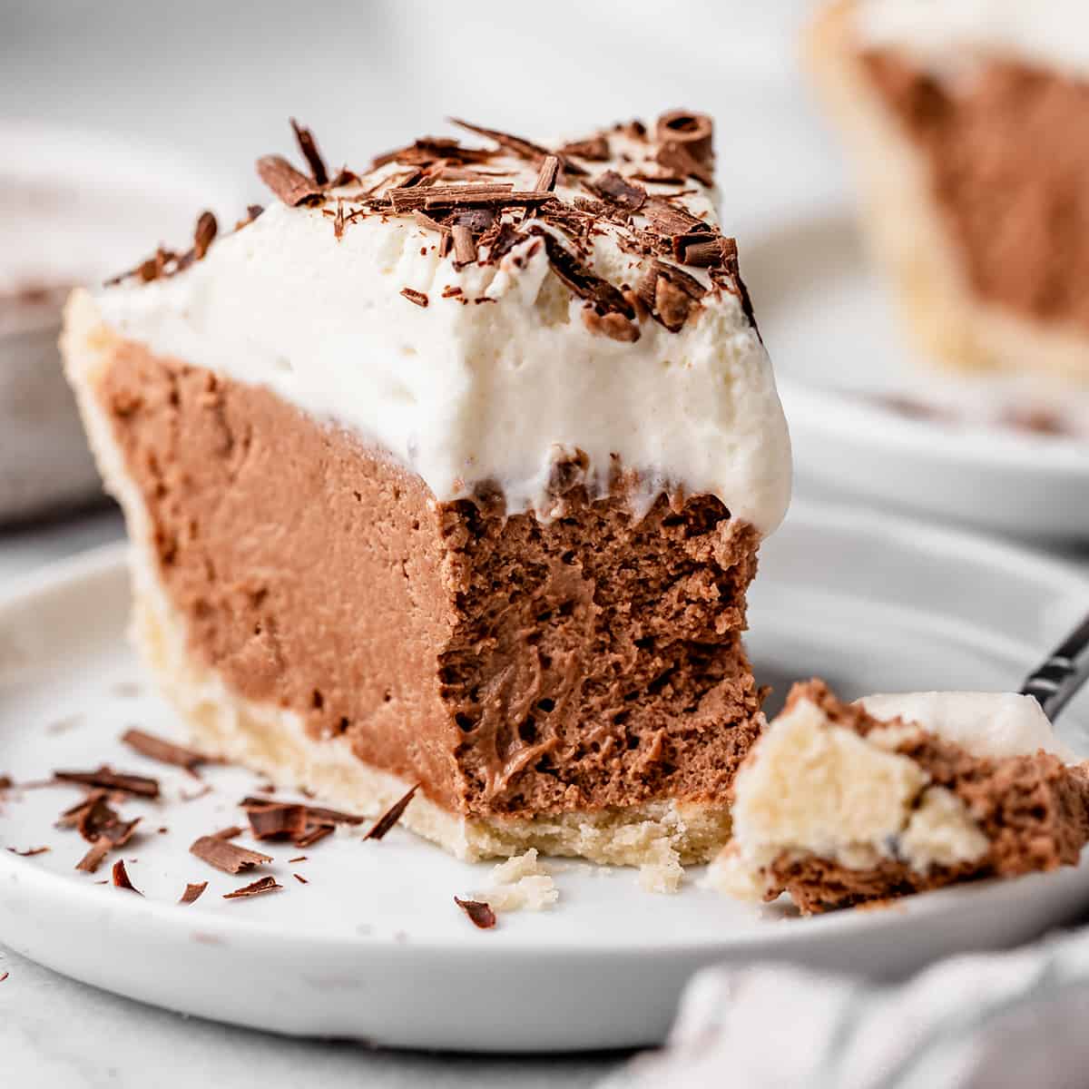 In this Homemade French Silk Pie recipe, a flaky pie crust is filled with a chocolate cream filling and topped with homemade whipped cream! This chocolate silk pie recipe is made from scratch and tastes even better than Baker's Square or Village Inn! #frenchsilkpie #frenchsilk #chocolatesilk French Silk Pie on a plate with a bite taken out of it