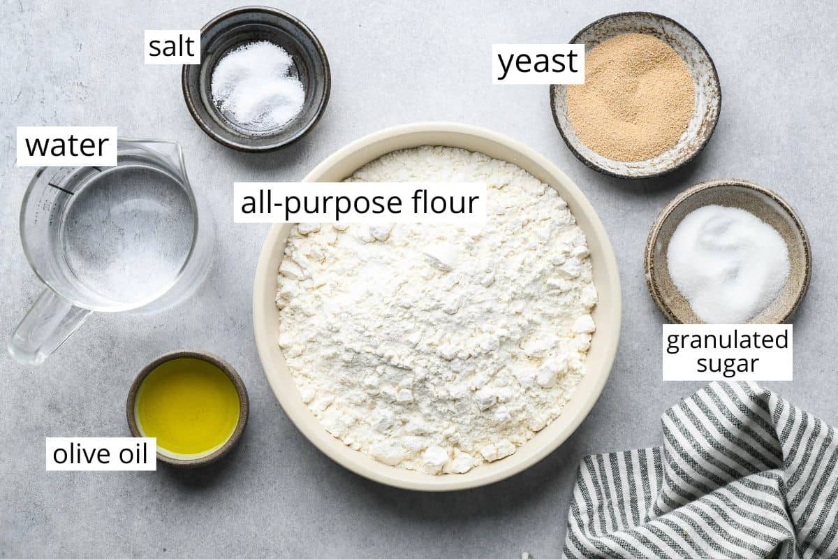 overhead photo of the labeled ingredients in this pizza dough recipe in bowls, flour, yeast, sugar, olive oil, water and salt