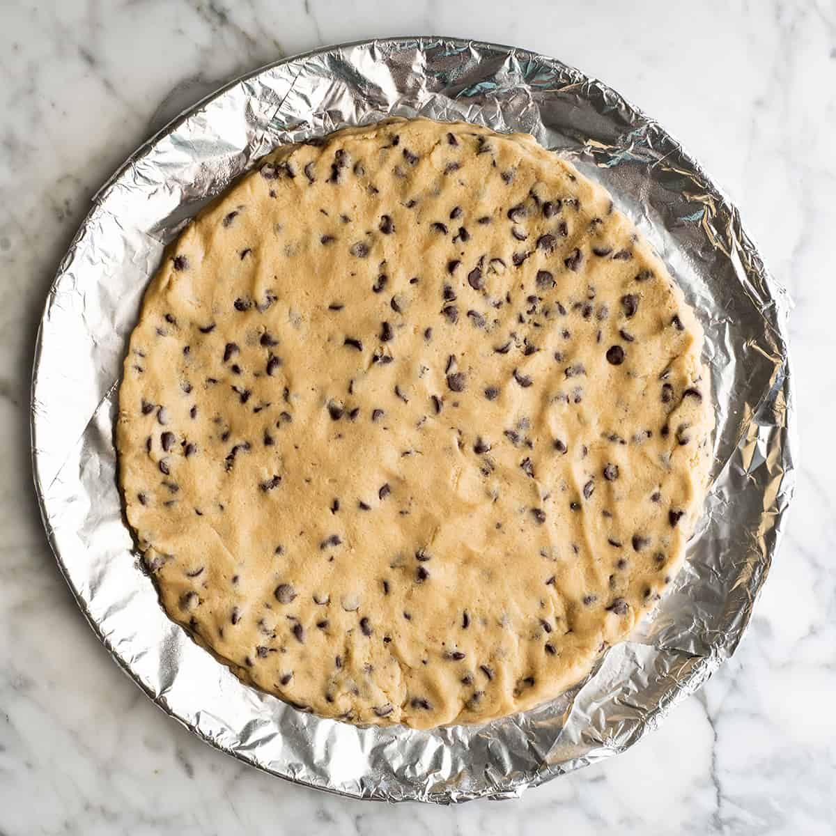 This chocolate chip cookie cake recipe is soft, chewy, slightly doughy and decorated with the best chocolate buttercream frosting. It's easy to make, large enough to feed a crowd, and even better than store-bought! chocolate chip cookie cake on a baking sheet before baking
