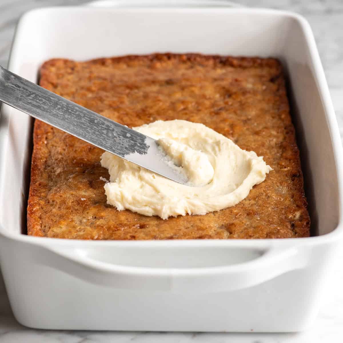 This is the best banana cake recipe ever! It's easy to make (no mixer required), moist, loaded with bananas and topped with a delicious cream cheese frosting. a metal spatula spreading cream cheese frosting on top of a banana cake in a white ceramic baking dish