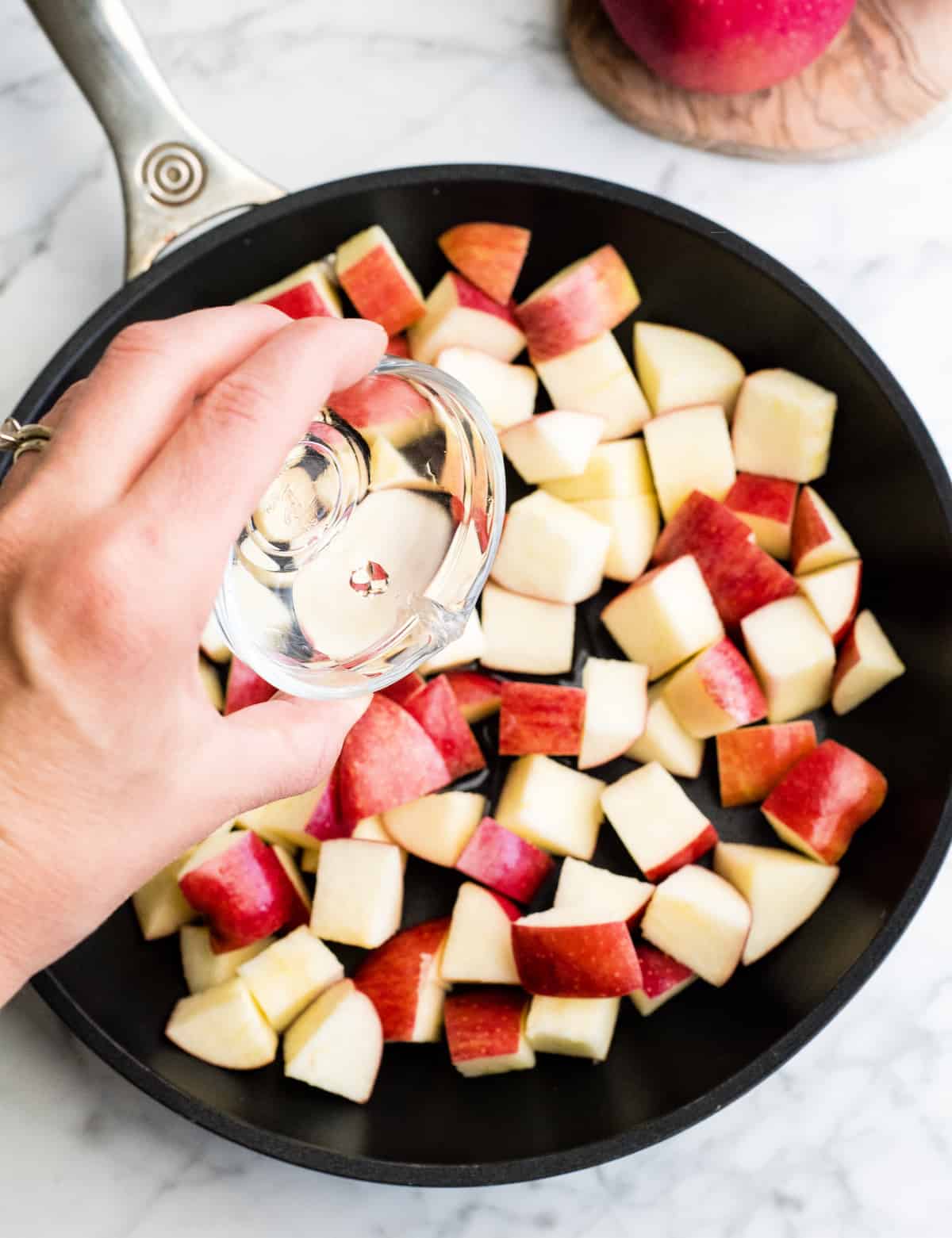 These Stovetop Sautéed Cinnamon Apples taste like a warm apple pie, but they come together in 5 minutes and are SO much healthier! This recipe is gluten-free, dairy-free, refined sugar free, vegan AND paleo! Perfect for breakfast, a snack, or dessert! #apples #stovetop #cinnamon #healthyrecipe #dessert #breakfast #snack #paleo #vegan #glutenfree #dairyfree #refinedsugarfree overhead view of a hand pouring water into diced apples in a nonstick fry pan showing how to make cinnamon apples