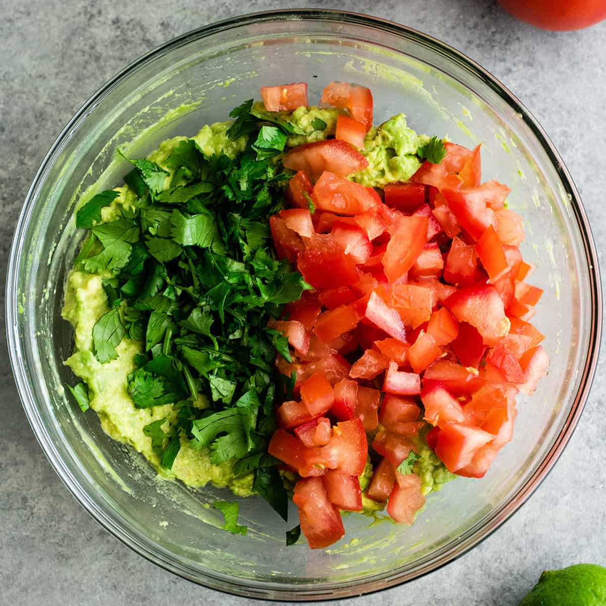 This quick & easy guacamole recipe is made with a handful of healthy ingredients and is ready in 5 minutes! This simple guacamole dip is our favorite appetizer and we always make it on taco nights! Watch the video to learn how to make this guacamole recipe! #guacamole #guacamolerecipe #tacos #appetizer #dip photo showing How to make Guacamole - adding cilantro, tomato and onion