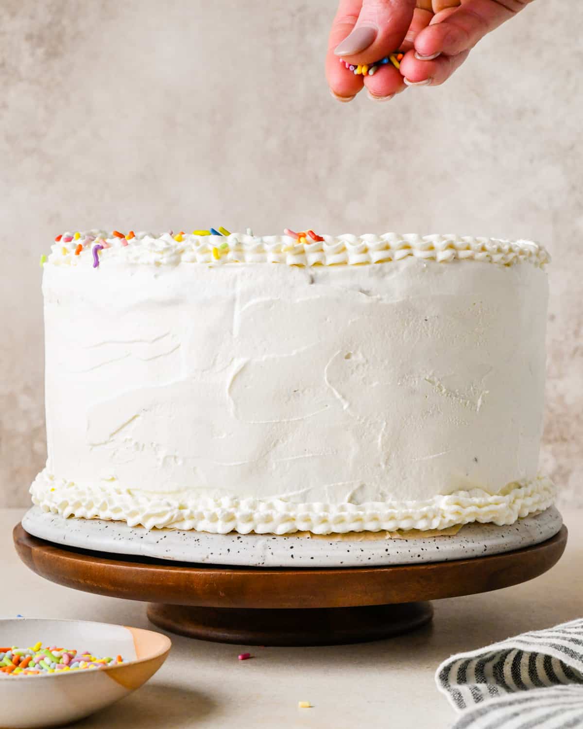 a hand sprinkling sprinkles over the whipped cream on an ice cream cake