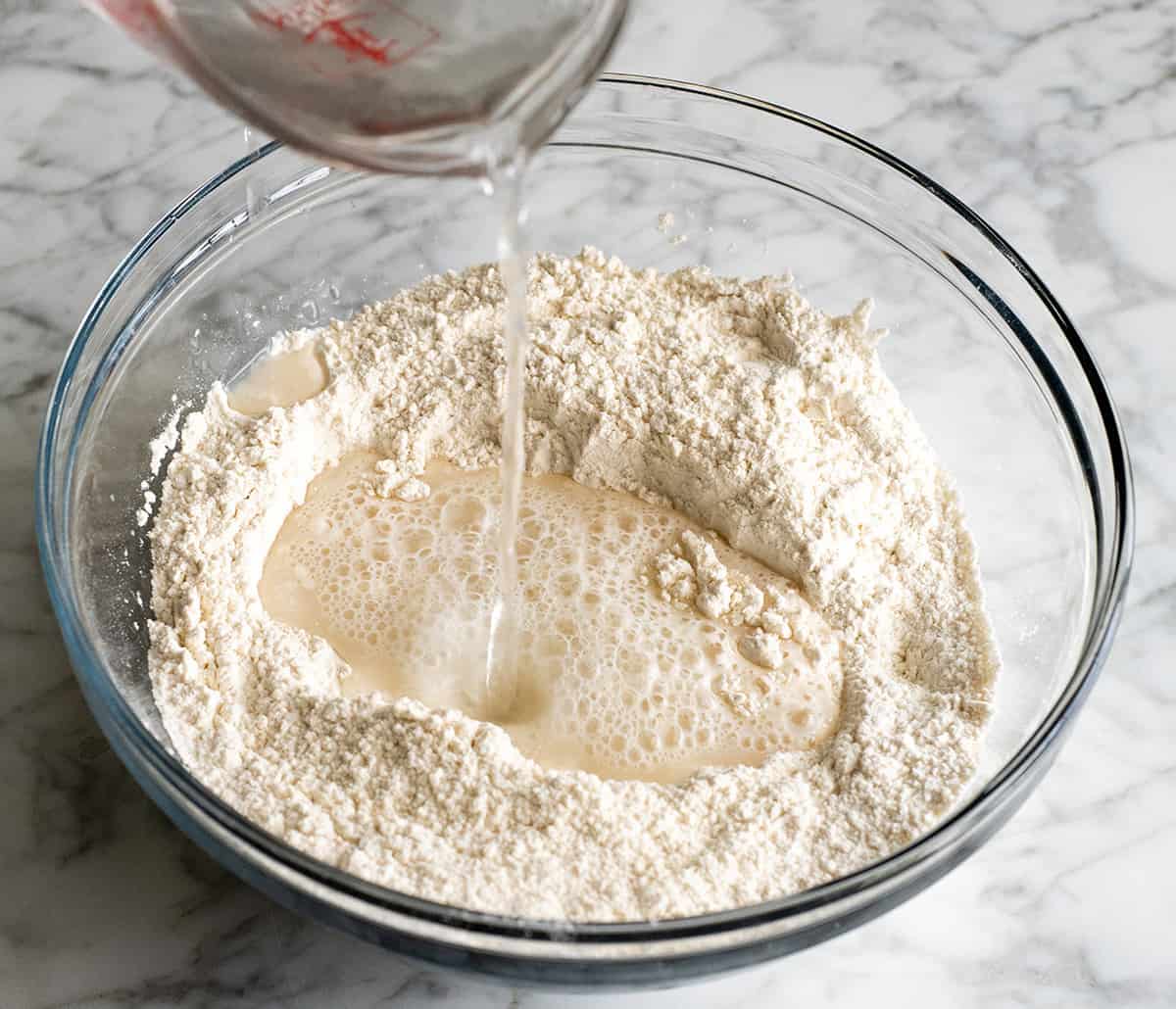 Easy Dutch Oven No Knead Bread recipe is made with only 4 ingredients and takes 5 minutes of prep time! If you want to make delicious homemade bread that looks & tastes like it came from a fancy bakery but actually takes very little effort, this dutch oven bread recipe is for you! front view of water being added showing how to make no knead bread