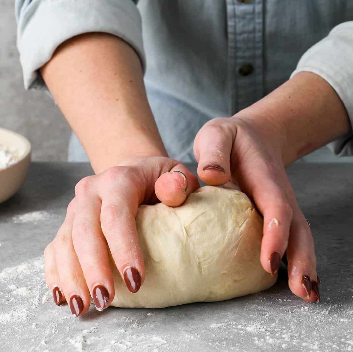hands kneading pizza dough on a gray floured surface with a bowl of flour nearby