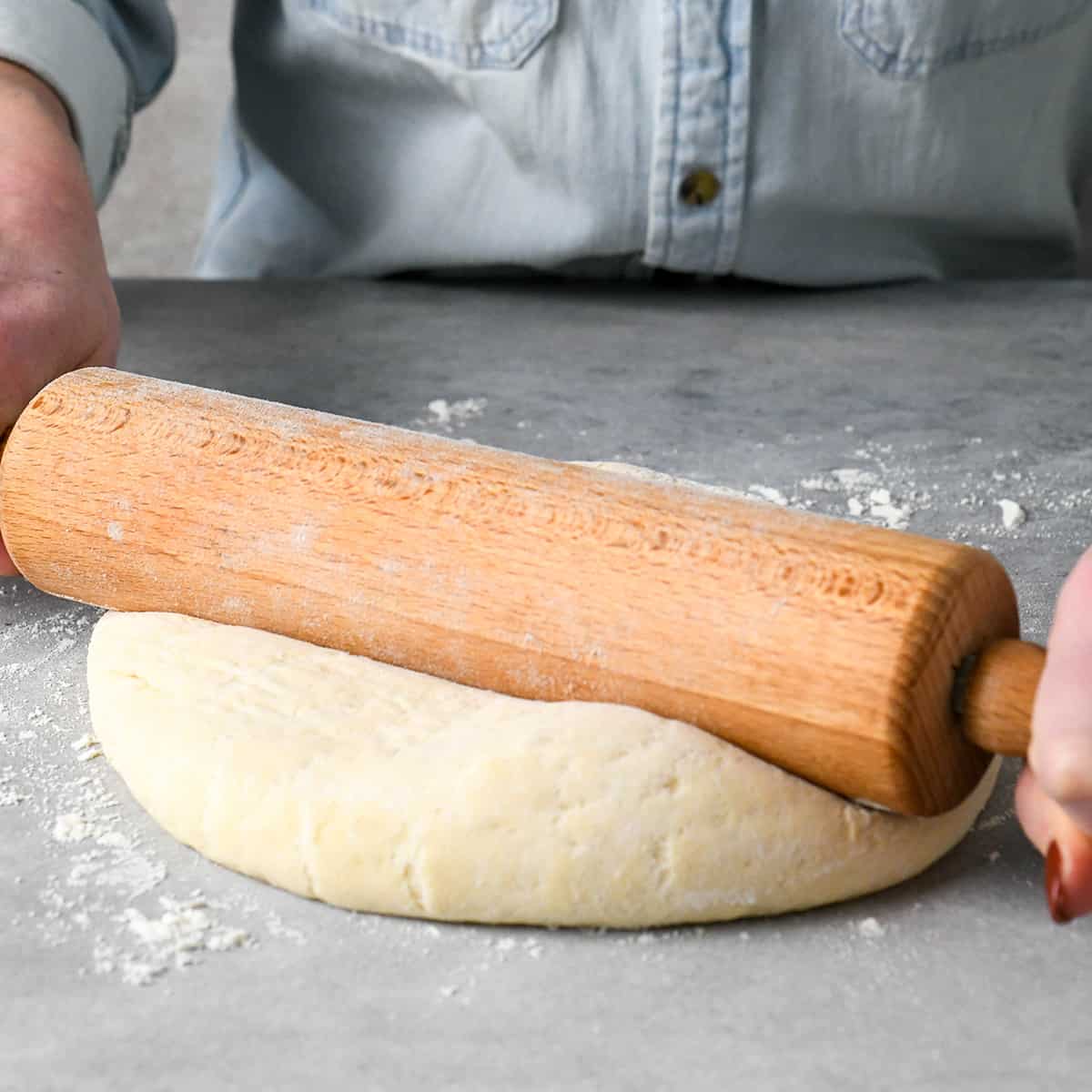 a woman rolling out pizza dough with a wooden rolling pin on a floured gray surface