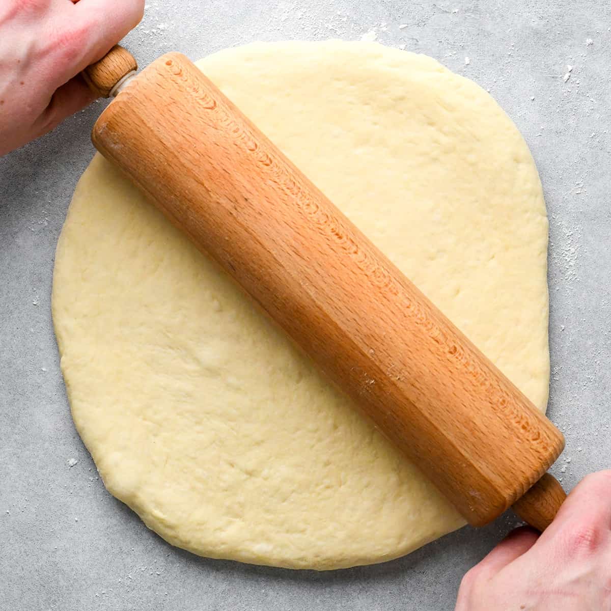 hands rolling out pizza dough with a wooden rolling pin on a floured gray surface